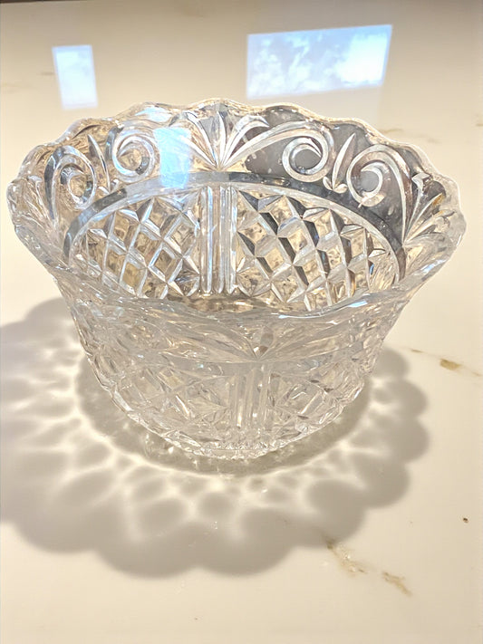 A luxury crystal serving bowl with intricate cut patterns and a scalloped rim sits on a light surface, reflecting soft light and casting decorative shadows. The detailed facets and clear glass create a sparkling effect, highlighting its elegant craftsmanship. The setting emphasizes a refined, timeless look ideal for serving or display in a kitchen, on a coffee table, or dining area.