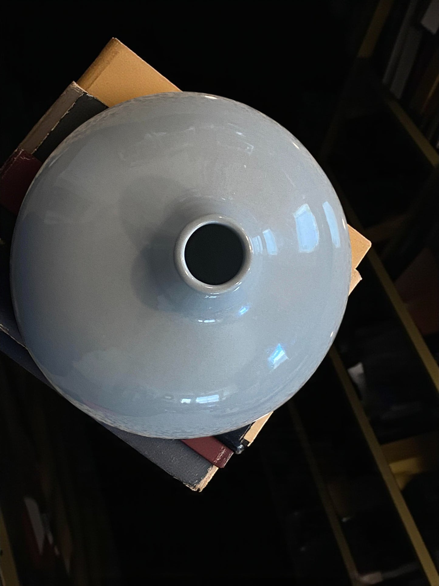 Close up of the top of a rounded ceramic vase with a smooth gray-blue glaze sits atop stacked books on a textured wood dresser. The minimalist design features a narrow neck and soft curves. It creates a calm, modern atmosphere, highlighting the vase as a simple, elegant focal point. Bohemian and rustic decor.