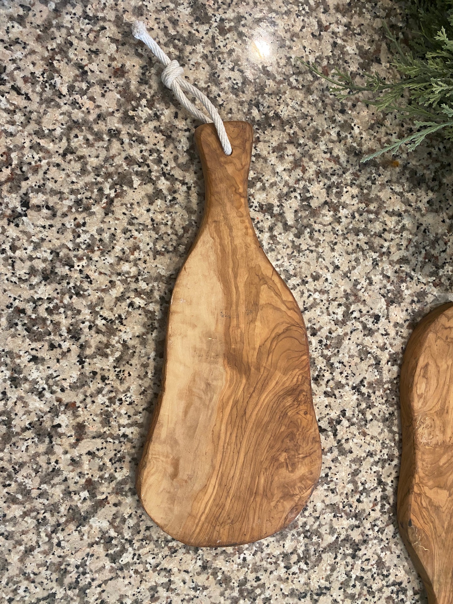 Live edge wooden cutting board with a organic, irregular shape is displayed on a granite countertop. One board lying flat with a rope on the handle to hang. A greenery garland with eucalyptus and berries sits in the background, emphasizing the boards’ natural wood grain and rustic, handcrafted look.