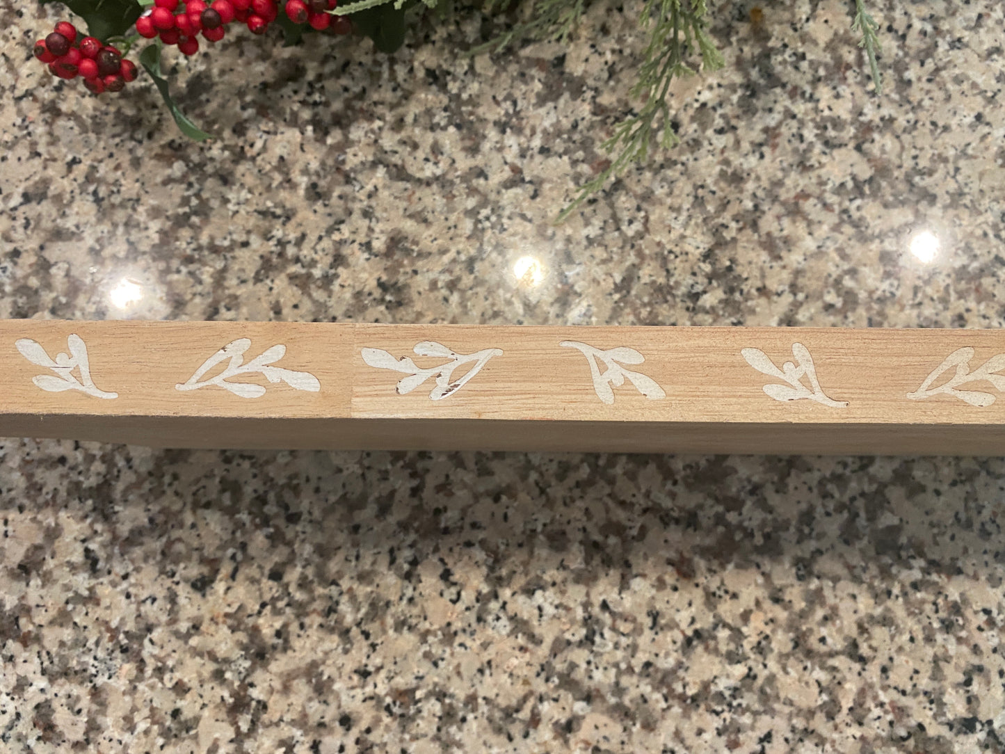A long rectangular wooden cutting board sits on a granite countertop, styled on a kitchen island. Behind it, a festive greenery garland with red berries adds a seasonal touch. The board’s natural wood grain, organic plant design on the sides, and elongated shape make it ideal for serving charcuterie or entertaining displays.