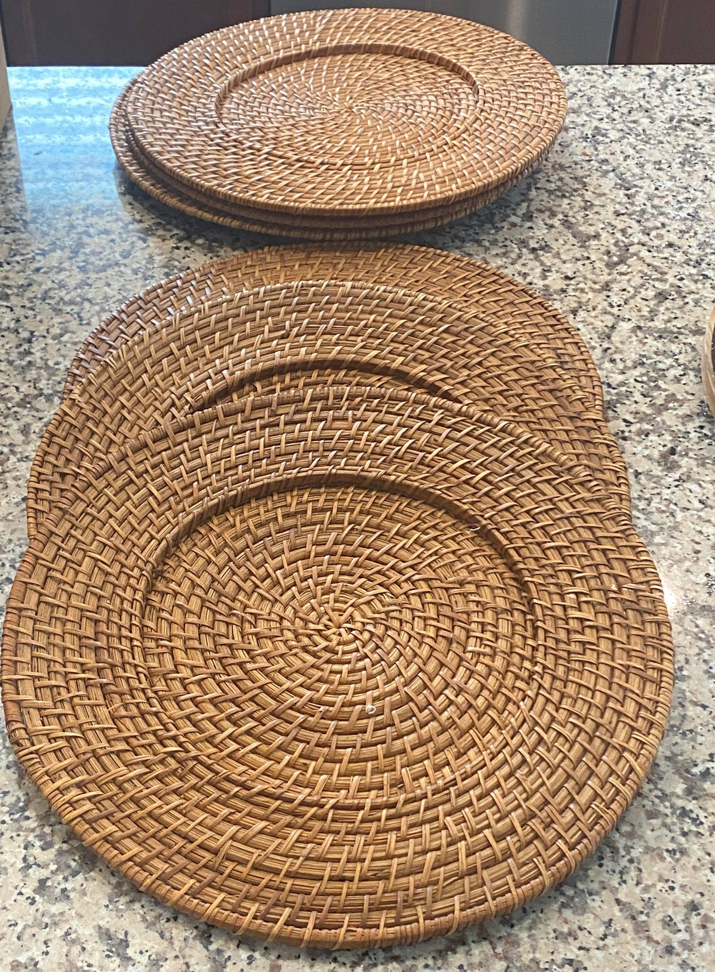 A set of six round wicker charger plates with a tightly woven rattan design is displayed on a speckled stone countertop. The plates feature a warm natural brown tone and a circular pattern that radiates from the center. Some chargers are stacked in the background while others are spread out in the foreground, highlighting their texture and handcrafted appearance. Great for bohemian, rustic, or bringing warmth to a modern dining table.