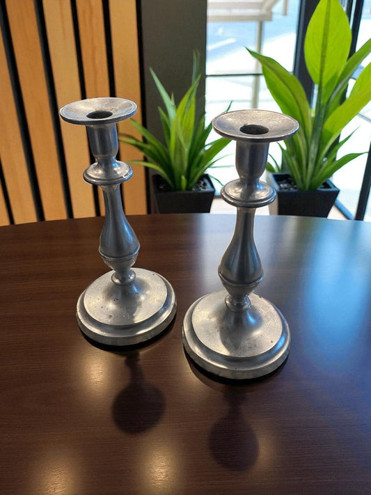 Two metal candle holders with a classic, sculptural design and aged silver finish sit on a dark wooden table. Behind them, green potted plants and a large window,  add a natural touch, while soft light highlights their smooth curves and subtle patina. The scene creates a warm, elegant, and timeless atmosphere with a blend of natural and refined elements.