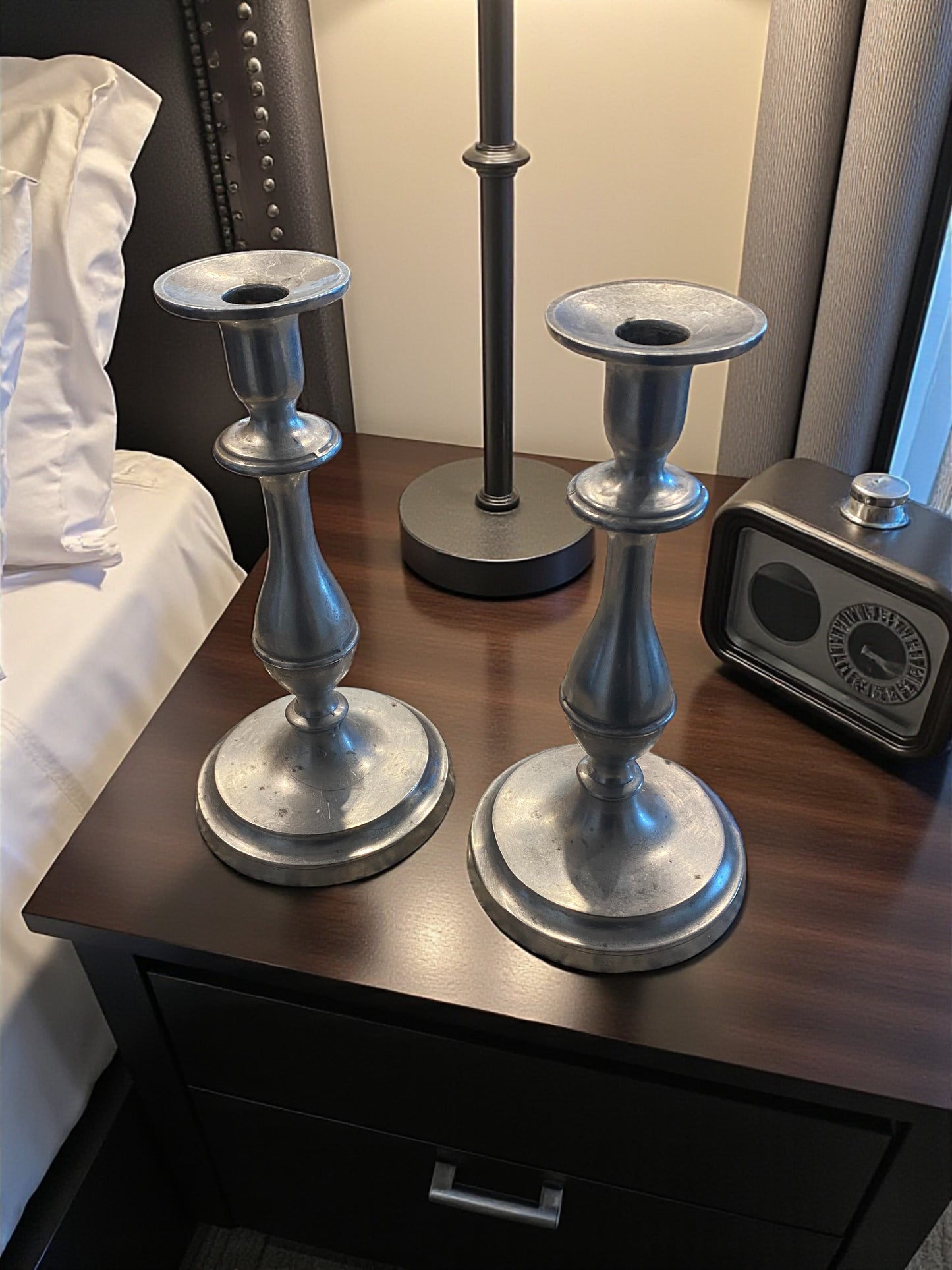 Two metal candle holders with a classic, sculptural design and aged silver finish sit on a dark wooden side table next to a bed. Behind them, a lamp and a clock,   while soft light highlights their smooth curves and subtle patina. The scene creates a warm, elegant, and timeless atmosphere with a blend of natural and refined elements.