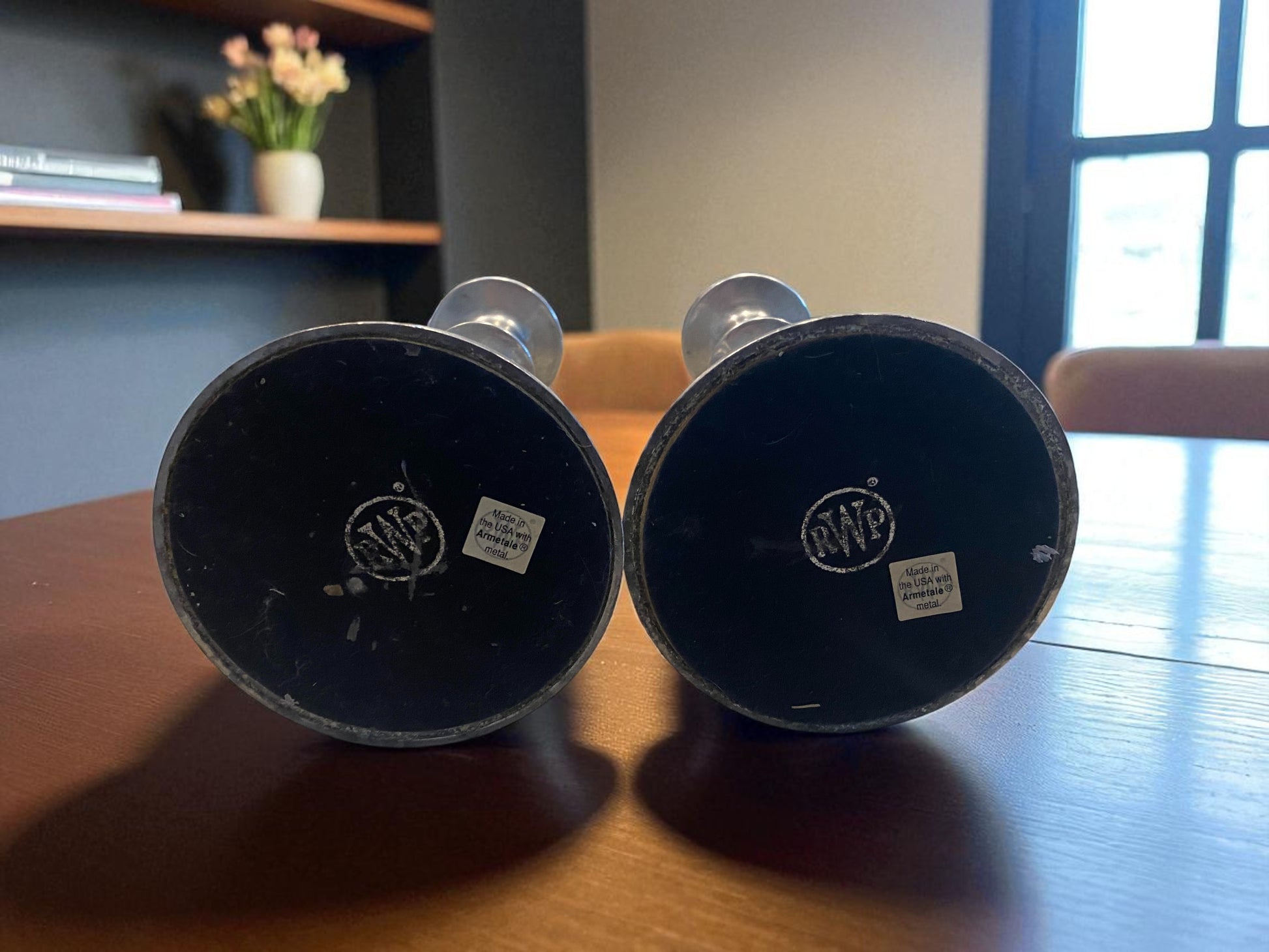 The bottom of two metal candle holders with a classic, sculptural design and aged silver finish sit on a dark wooden table. Showing the makers mark.  Behind them, green potted plants and a large window,  add a natural touch, while soft light highlights their smooth curves and subtle patina. The scene creates a warm, elegant, and timeless atmosphere with a blend of natural and refined elements.