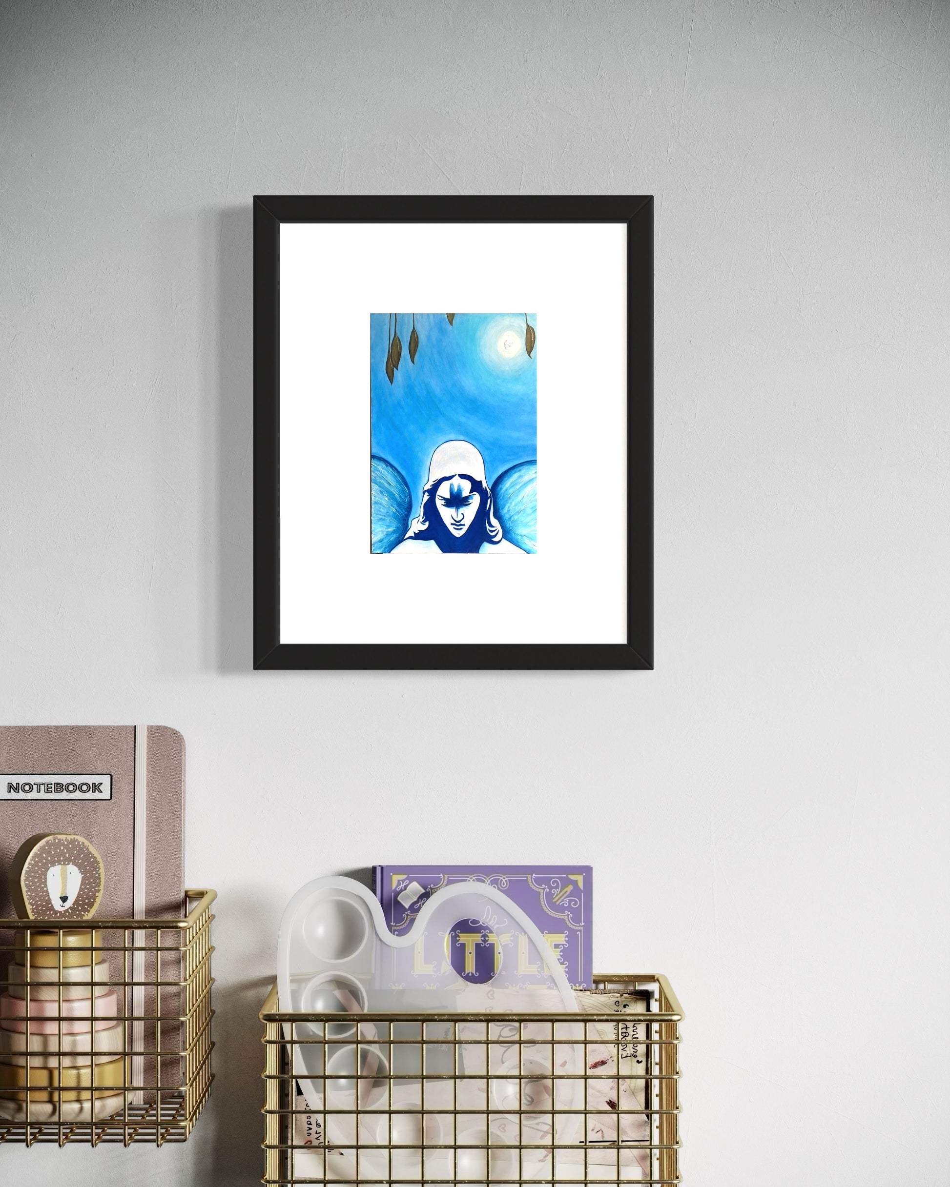 A  framed blue-toned art print of a angel beneath a glowing moon, surrounded by soft gradients and subtle details. hung on a white wall.  Hung above two wire bins that have a note book and painters palette within them.  Creating a calm, balanced, reflective, and contemporary atmosphere.