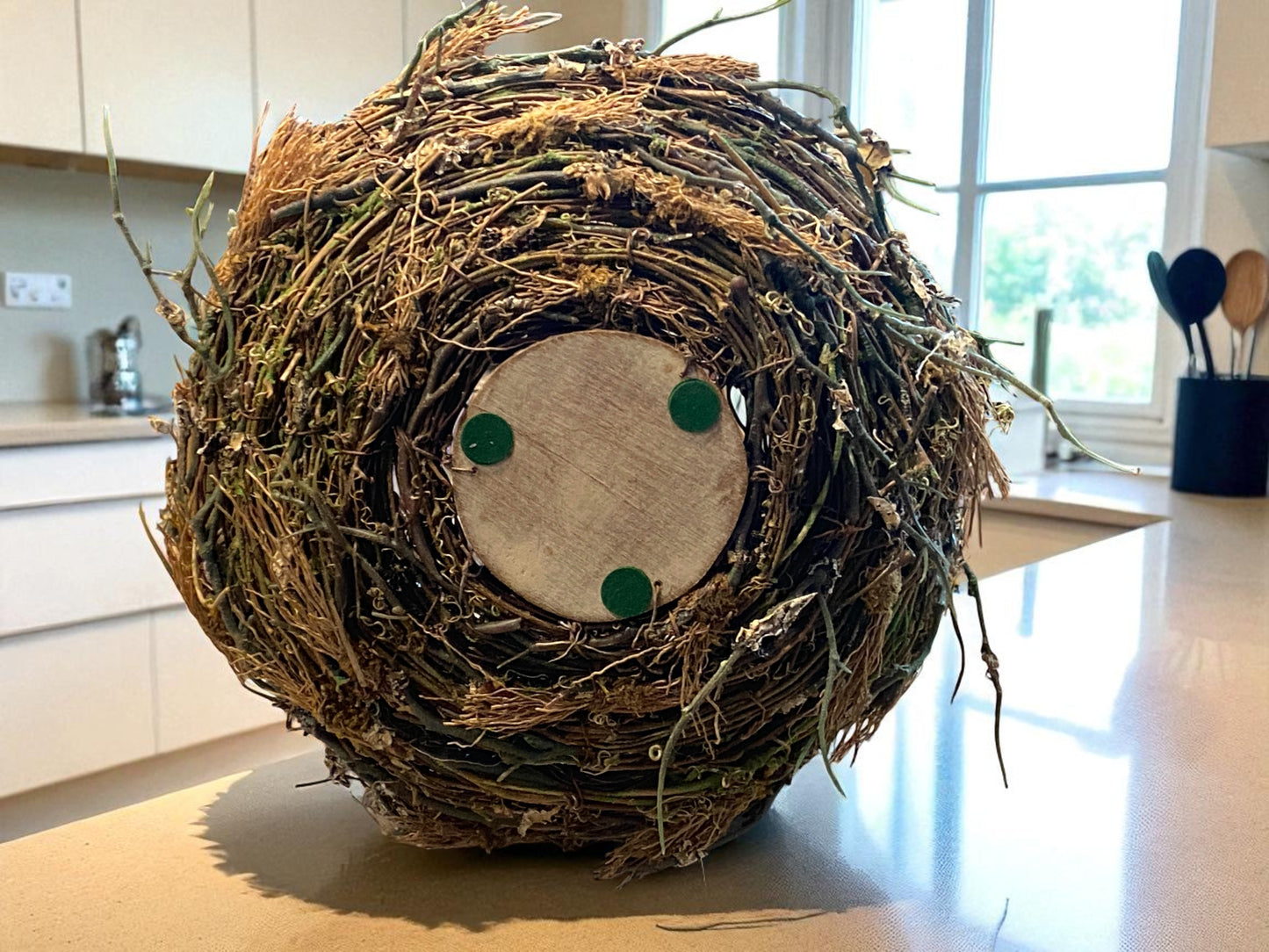 Close-up of the bottom view of the organic twig and moss texture of bird’s nest planter made of intertwined mixture of artificial twigs and natural fibers sits on a white granite kitchen countertop. A clean white kitchen and window in the background.  The organic, textured design features earthy brown and green tones, adding a rustic, nature-inspired touch.   Great for any kitchen, bathroom, sun room, foyer, dining area, living room or office.  