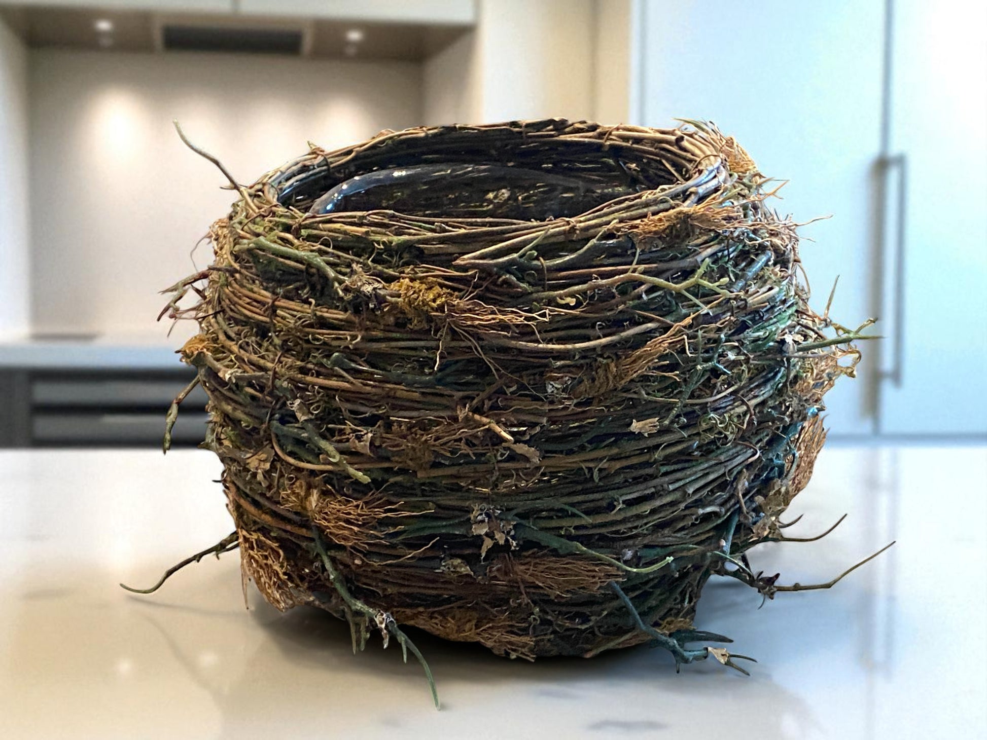 A bird’s nest-style planter made of intertwined mixture of artificial twigs and natural fibers sits on a granite counter top. The organic, textured design features earthy brown and green tones, adding a rustic, nature-inspired touch. The background of a white modern kitchen creates a warm, balanced contrast between natural materials and clean, contemporary surfaces.  Great for any room of a modern home - kitchen, bathroom, office, den, or foyer.