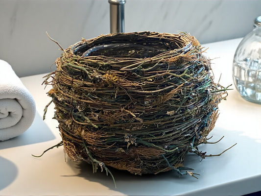 A bird’s nest-style planter made of intertwined mixture of artificial twigs and natural fibers sits on a bathroom countertop, with a towel and glass container next to it. The organic, textured design features earthy brown and green tones, adding a rustic, nature-inspired touch. The soft background of a white/gray marbled modern bathroom creates a warm, balanced contrast between natural materials and clean, contemporary surfaces.  Great for any room of a modern home - kitchen, bathroom, office, den, or foyer