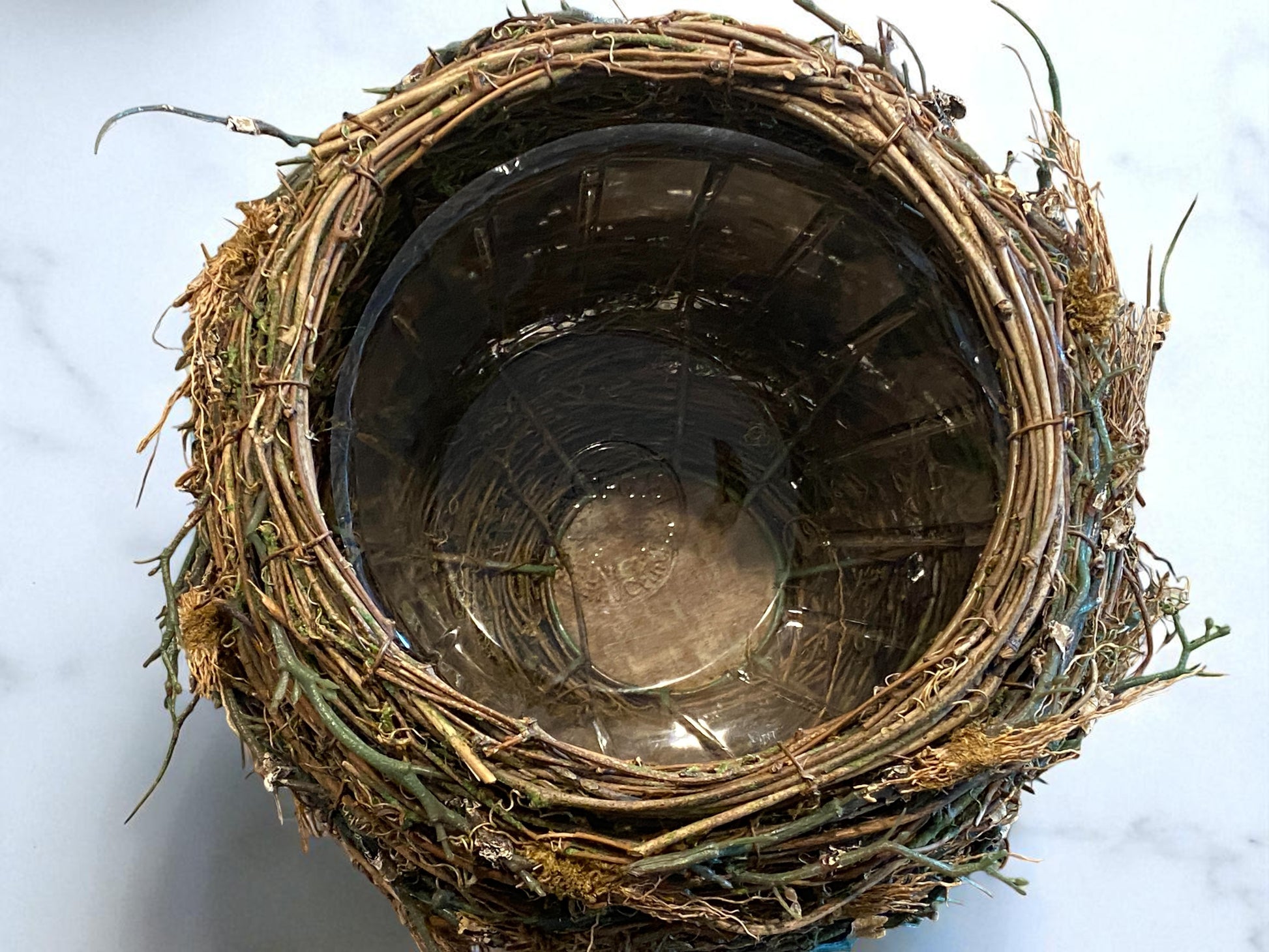 Close-up of the top view of the organic twig and moss texture of bird’s nest planter made of intertwined mixture of artificial twigs and natural fibers sits on a white granite countertop. The organic, textured design features earthy brown and green tones, adding a rustic, nature-inspired touch.   Great for any kitchen, bathroom, sun room, foyer, dining area, living room or office.  