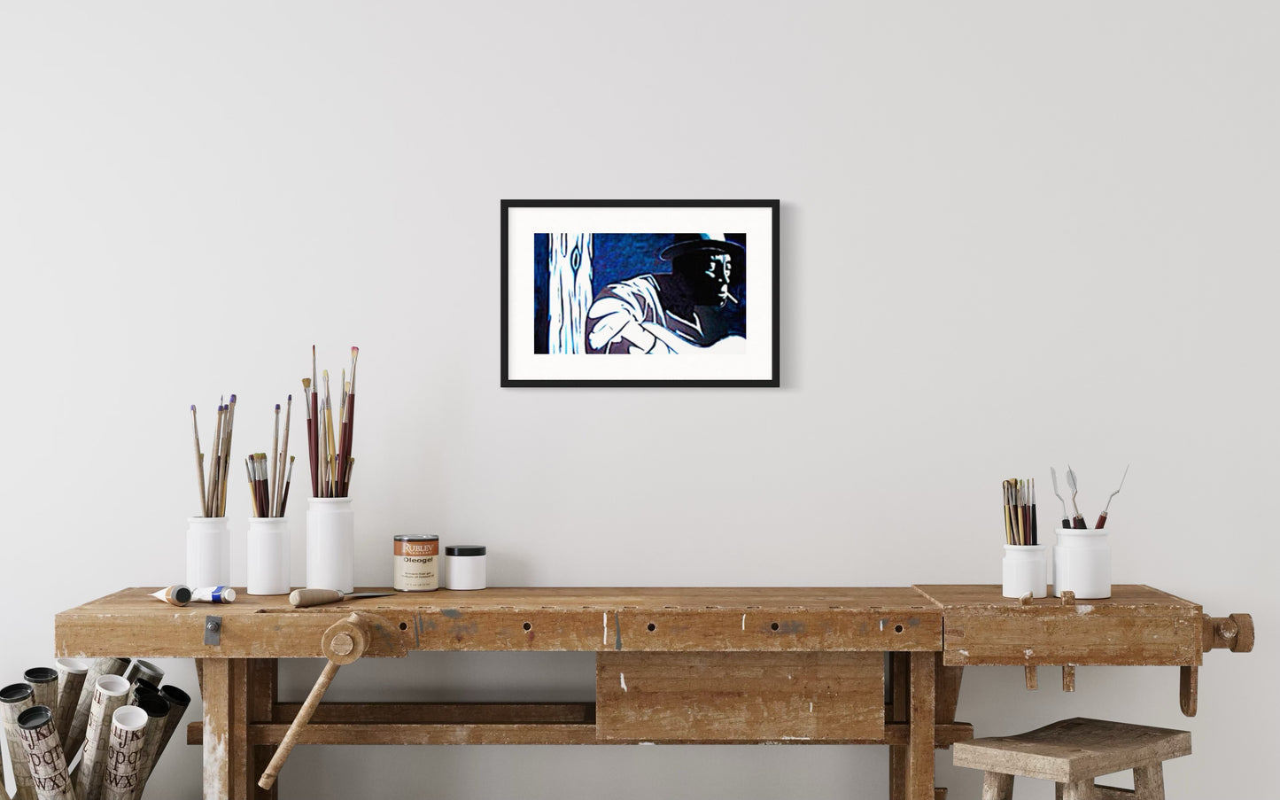 A stylish interior scene featuring a framed blues player-inspired art print of a musician in dark smokey room, Mississippi John Hurt.   Monochromatic style hanging above an artist work bench displaying art brushes, palette knives, and paints.  Together giving the space a warm, creative, and music-inspired atmosphere.