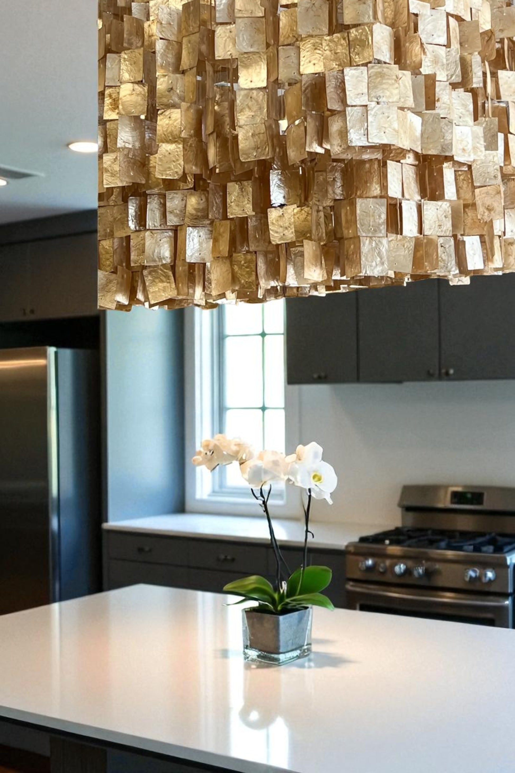 A unique capiz shell chandelier with layered, translucent champaign/golden tiles hangs above a granite kitchen island, casting a warm, soft glow. Below, a simple floral arrangement sits at the center, while dark gray kitchen cabinets and appliances and a nearby window create contrast. The textured fixture adds elegance and visual interest, creating a modern yet organic atmosphere.  A focal point for any living room, kitchen, dining area, or foyer.  