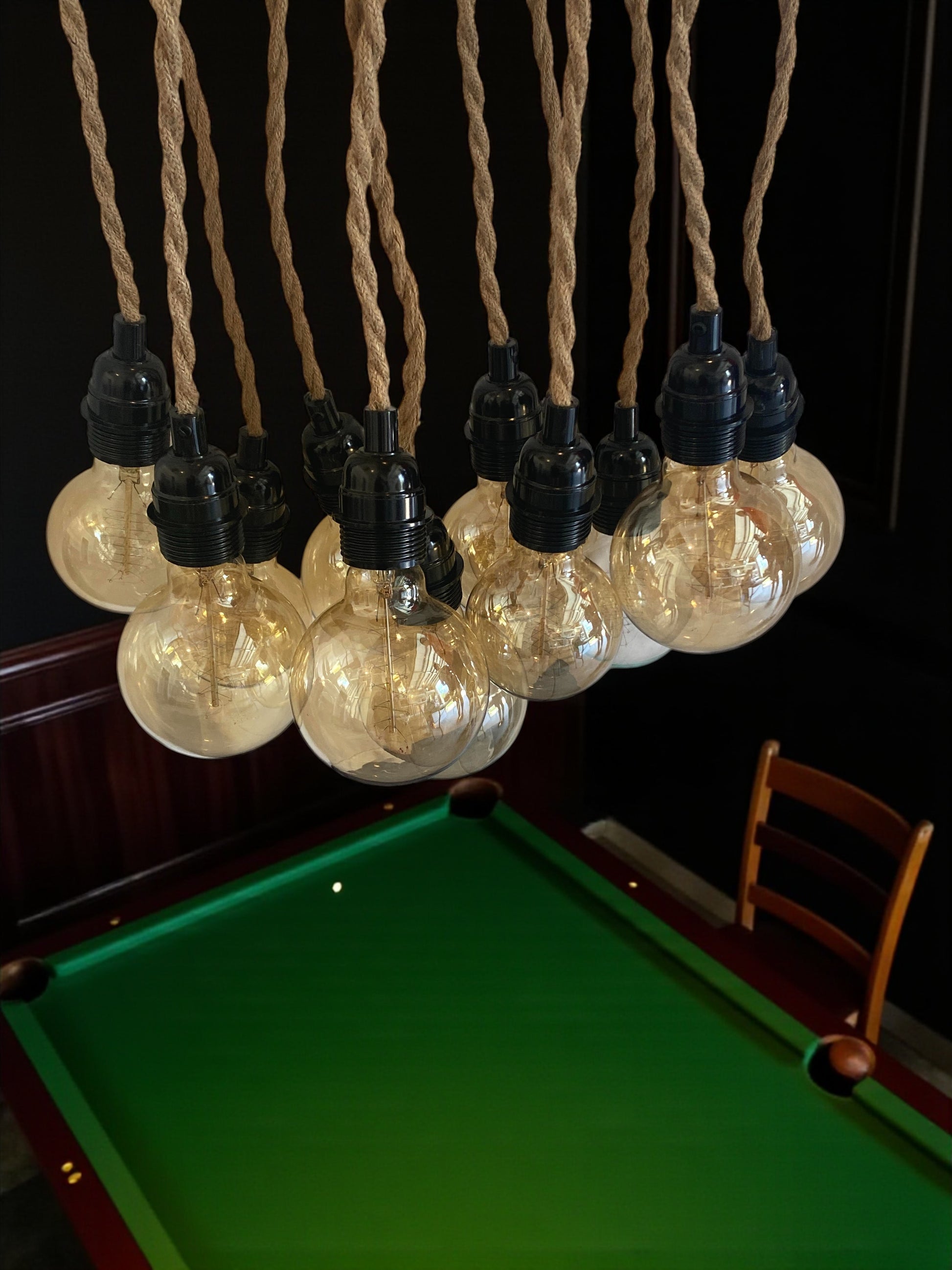 A unique, vintage-inspired chandelier made from a Chevy hubcap as ceiling cover and is mounted to a ceiling overlooking a pool table, game room with multiple twisted rope electrical cords hanging down. Each cord holds an exposed extra large Edison bulb with a warm amber glow. The fixture has an industrial, rustic look and is shown in a modern interior space, highlighting its unique design and vintage automotive influence.