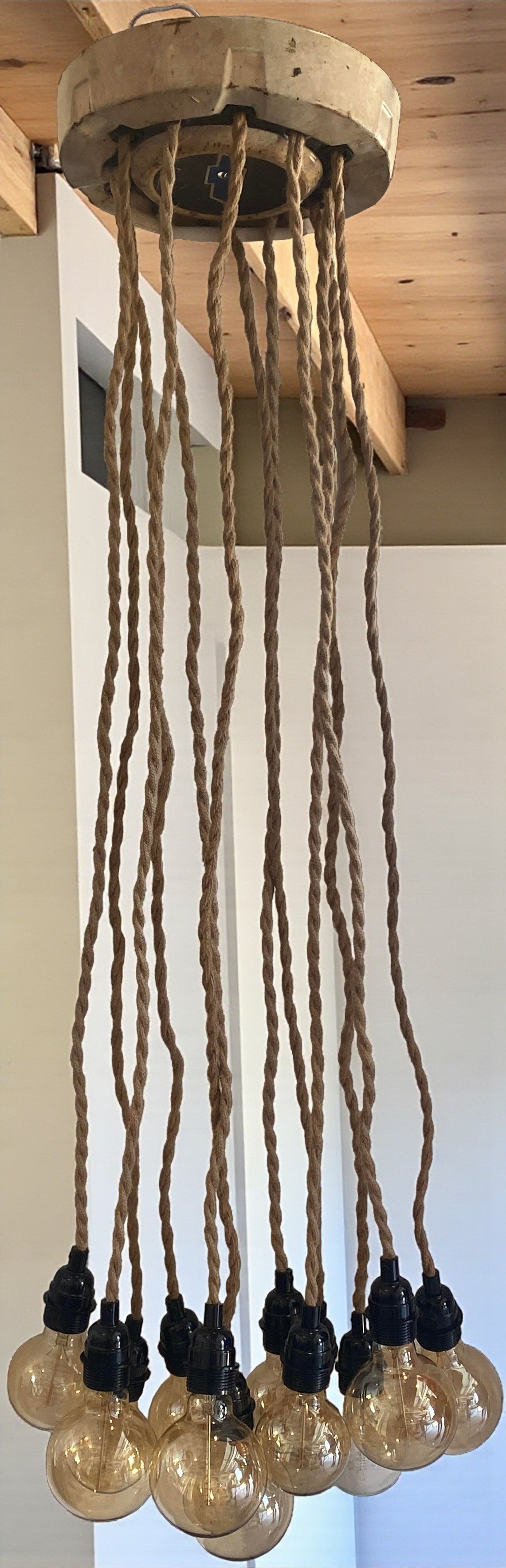 A unique, vintage-inspired chandelier made from a Chevy hubcap as ceiling cover and is mounted to a wood ceiling, with multiple twisted rope electrical cords hanging down. Each cord holds an exposed Edison bulb with a warm amber glow. The fixture has an industrial, rustic look and is shown in a modern interior space, highlighting its unique design and vintage automotive influence.