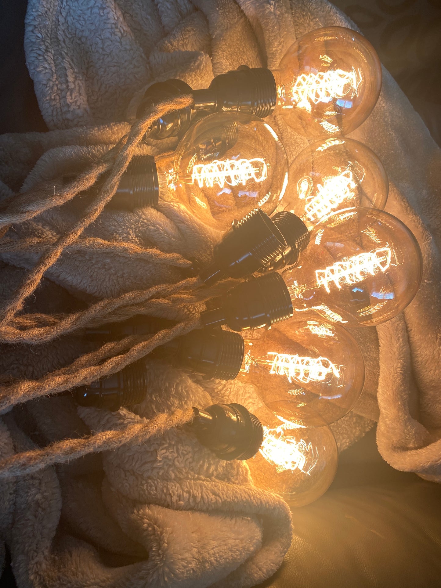 Warm ambient Edison bulbs that are at the end of a unique, vintage-inspired chandelier/pendant light made from a Chevy hubcap as ceiling cover and is mounted to a wood ceiling, with multiple twisted rope electrical cords hanging down. Each cord holds an exposed Edison bulb with a warm amber glow. The fixture has an industrial, rustic look and is shown in a modern interior space, highlighting its unique design and vintage automotive influence.