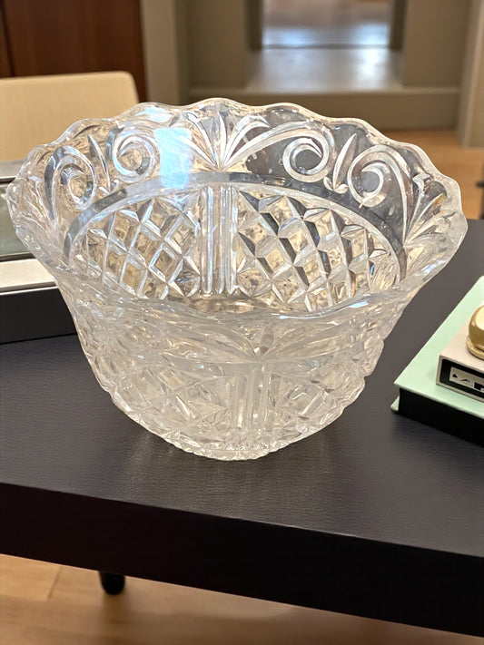 A luxury crystal serving bowl with intricate cut patterns and a scalloped rim sits on a light surface, reflecting soft light and casting decorative shadows. The detailed facets and clear glass create a sparkling effect, highlighting its elegant craftsmanship. The setting emphasizes a refined, timeless look ideal for serving or display in a kitchen, on a coffee table, or dining area.