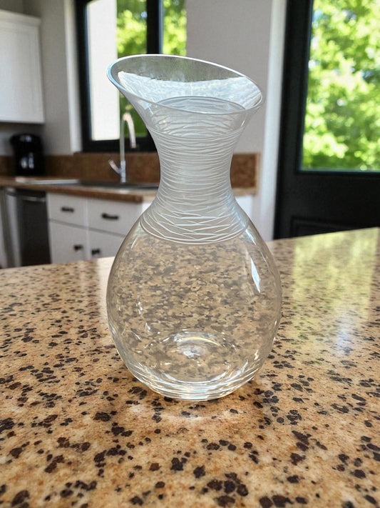 A clear glass Dansk decanter with a rounded base and flared, sculptural neck sits on a speckled stone countertop in a bright kitchen. The neck features delicate etched lines, adding texture and visual interest. Natural light from nearby windows highlights its smooth curves, creating a clean, modern, and elegant atmosphere suitable for serving or display.