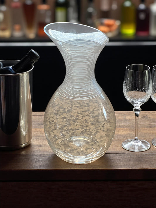 A clear glass Dansk decanter with a rounded base and flared, sculptural neck sits on dark wood bar countertop. The neck features delicate etched lines, adding texture and visual interest. Wine glasses and wine chiller next to it with liquor bottles on the opposite bar counter in the background.  Its smooth curves, creating a clean, modern, and elegant atmosphere suitable for serving or display.