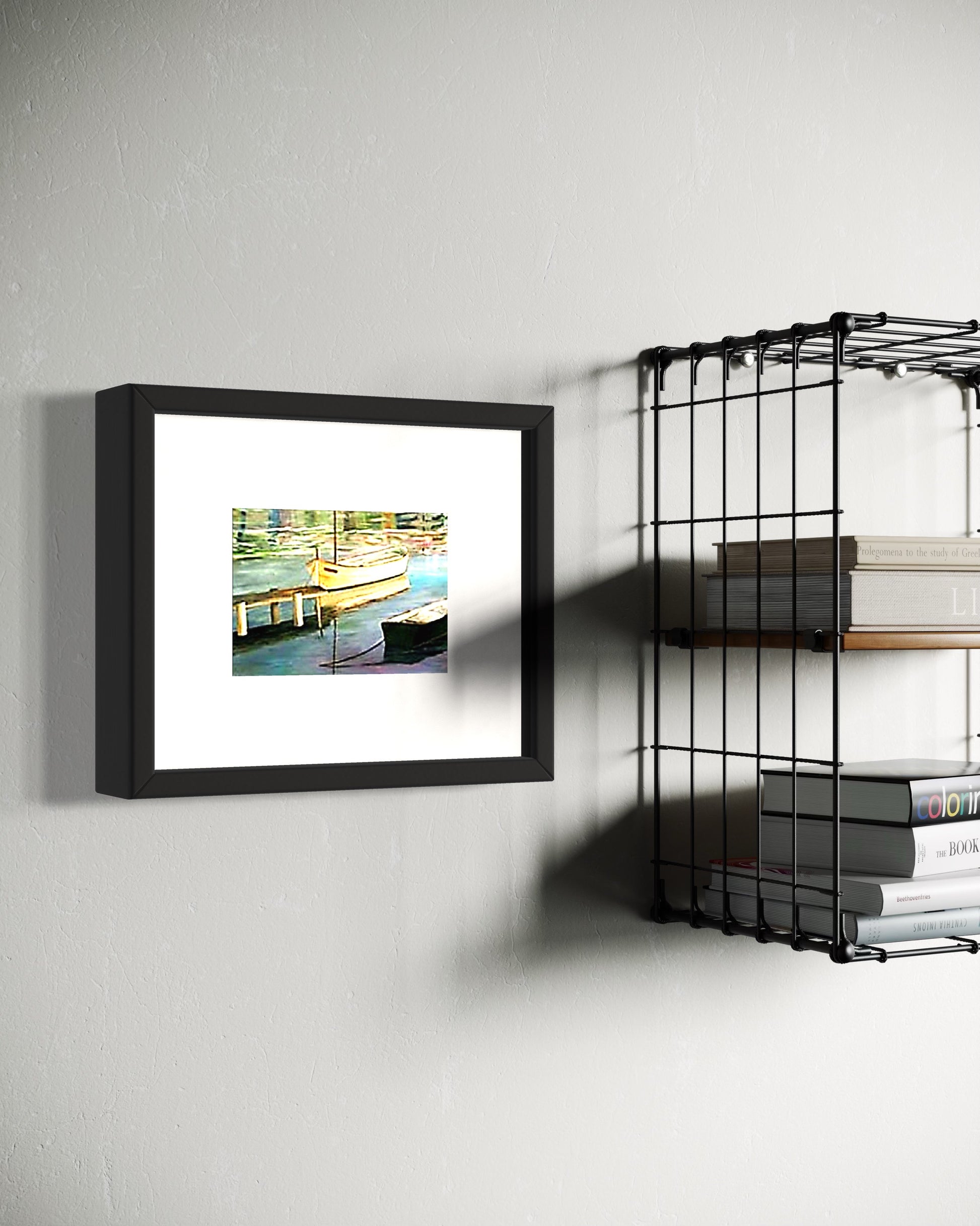 A framed coastal art print of a dock with a small boat reflected in calm water hangs on a white wall.  Next to industrial wire shelves with books.  The scene combines a warm sea side print with industrial shelves, creating a calm, classic atmosphere with a subtle coastal influence.