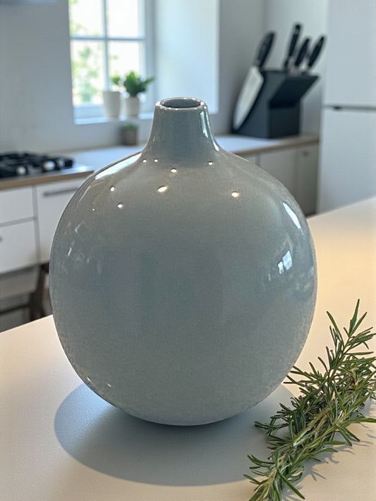 A rounded ceramic vase with a smooth gray-blue glaze sits atop a white stone kitchen island.  The minimalist design features a narrow neck and soft curves. In the background, a white modern kitchen with plants in the window creates a calm, modern atmosphere, highlighting the vase as a simple, elegant focal point.  Bohemian and rustic decor.  