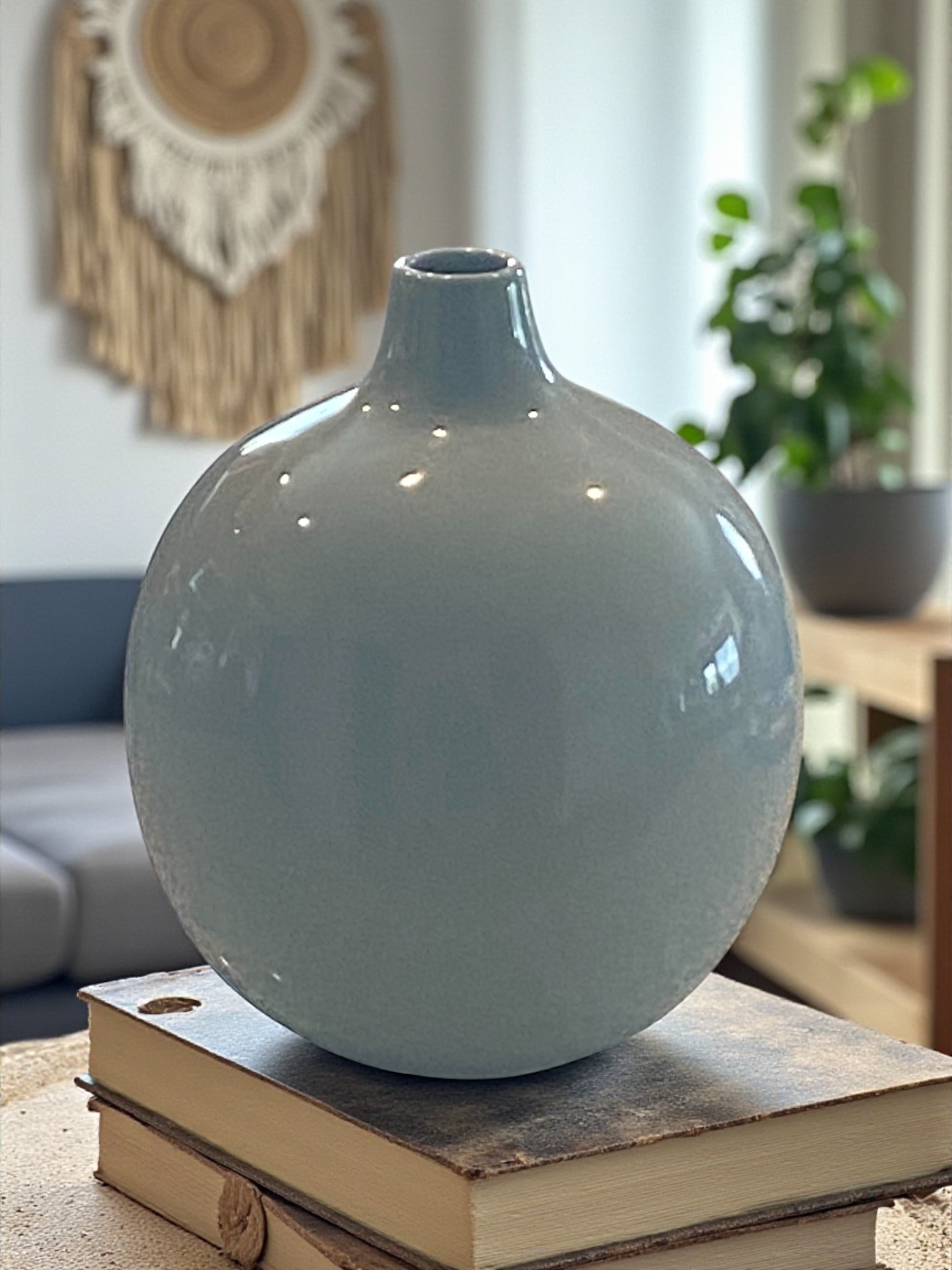 A rounded ceramic vase with a smooth gray-blue glaze sits atop stacked books on a textured wood coffee table. The minimalist design features a narrow neck and soft curves. In the background, a cozy living space with plants and woven wall decor creates a calm, modern atmosphere, highlighting the vase as a simple, elegant focal point.  Bohemian and rustic decor.  
