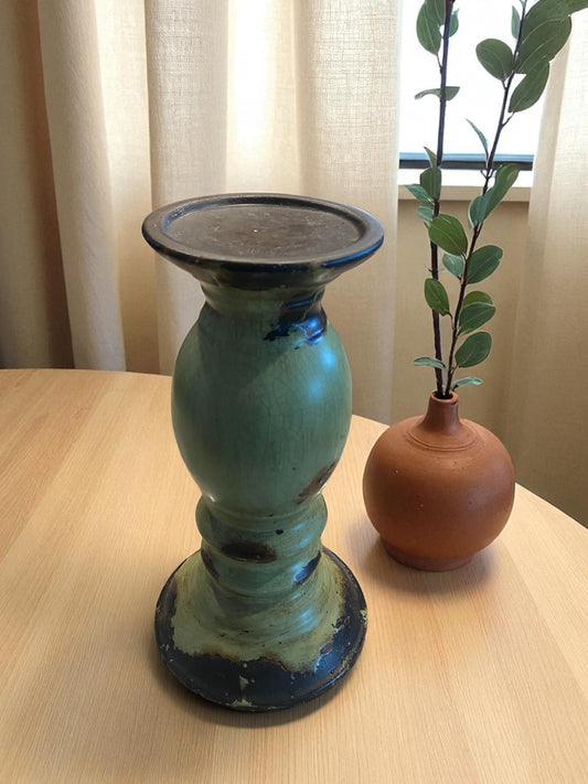 A large rustic green pottery candle holder with a distressed glaze and dark accents stands on a wooden table. Its sculptural shape features a wide top and sturdy base. Beside it, a small terracotta vase with a leafy stem adds contrast. Soft natural light and neutral curtains coming from a window create a warm, earthy, and inviting atmosphere.  Rustic or bohemian decor.  