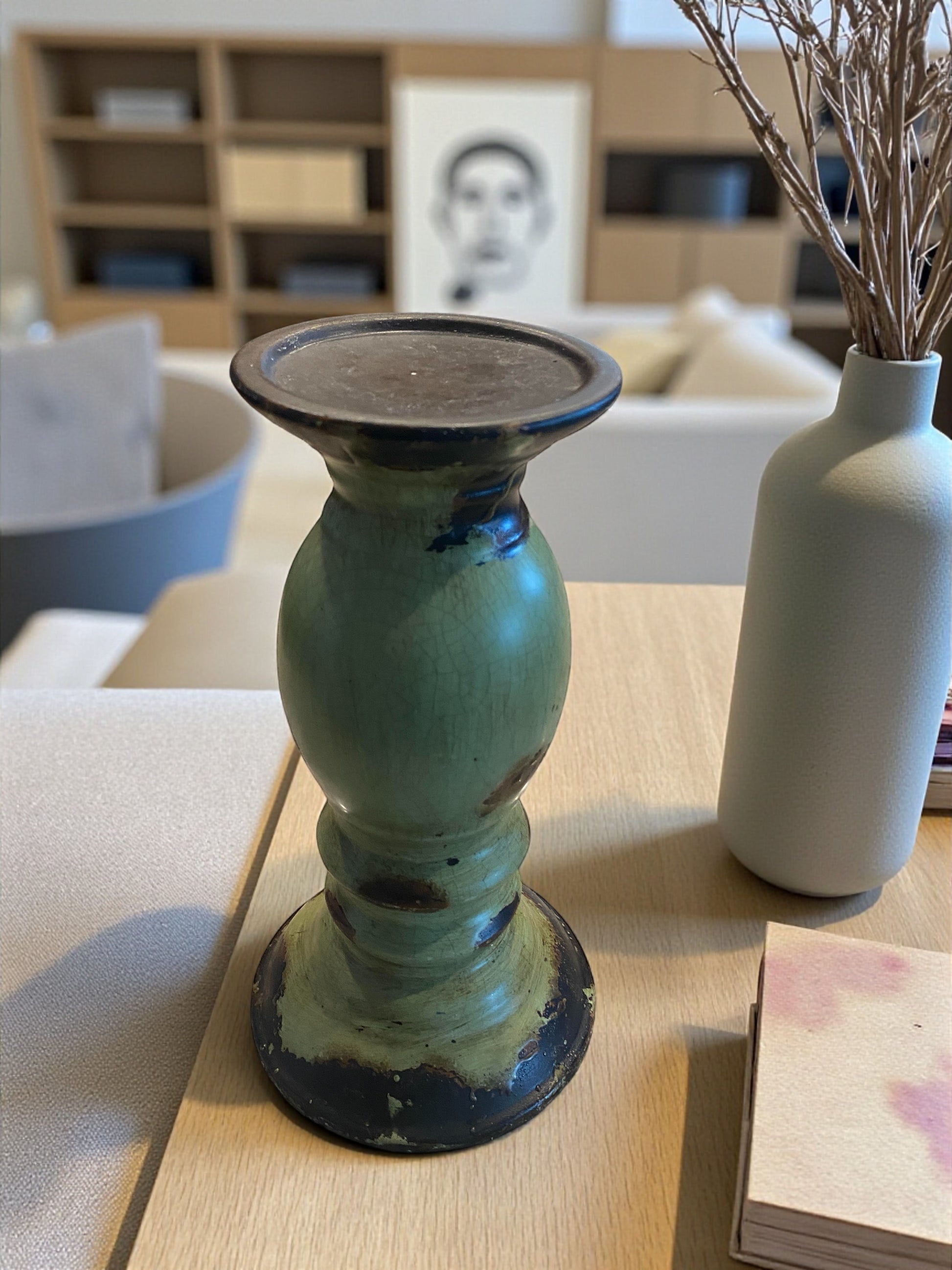 A large rustic green pottery candle holder with a distressed glaze and dark accents stands on a wooden table. Its sculptural shape features a wide top and sturdy base. Beside it, a small white vase with dried stems adds contrast. Soft natural light and neutral wood entertainment center, art, and couch in the background create a warm, earthy, and inviting atmosphere.  Rustic or bohemian decor.  