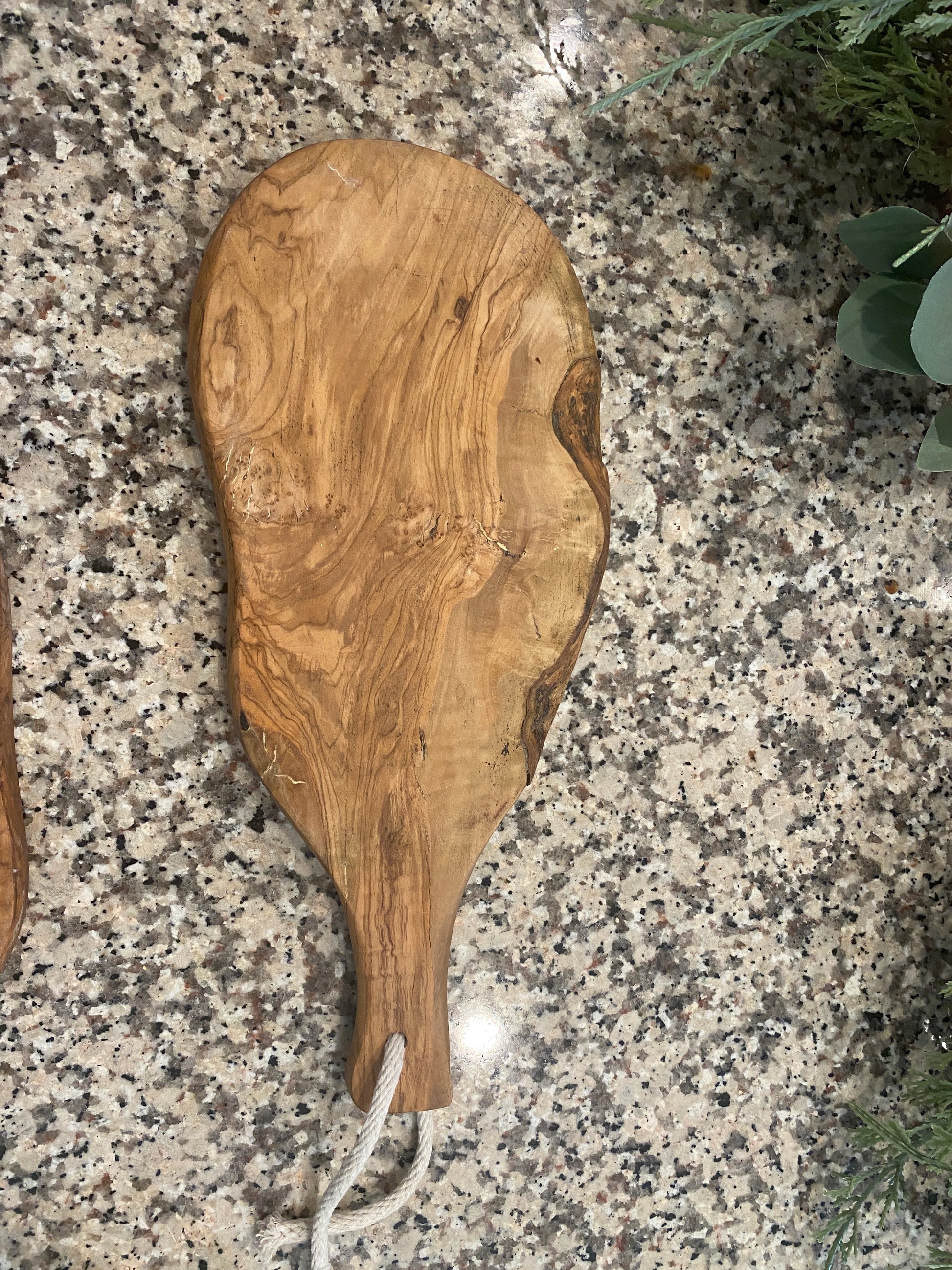 Live edge wooden cutting board with a organic, irregular shape is displayed on a granite countertop. One board lying flat with a rope on the handle to hang. A greenery garland with eucalyptus and berries sits in the background, emphasizing the boards’ natural wood grain and rustic, handcrafted look.