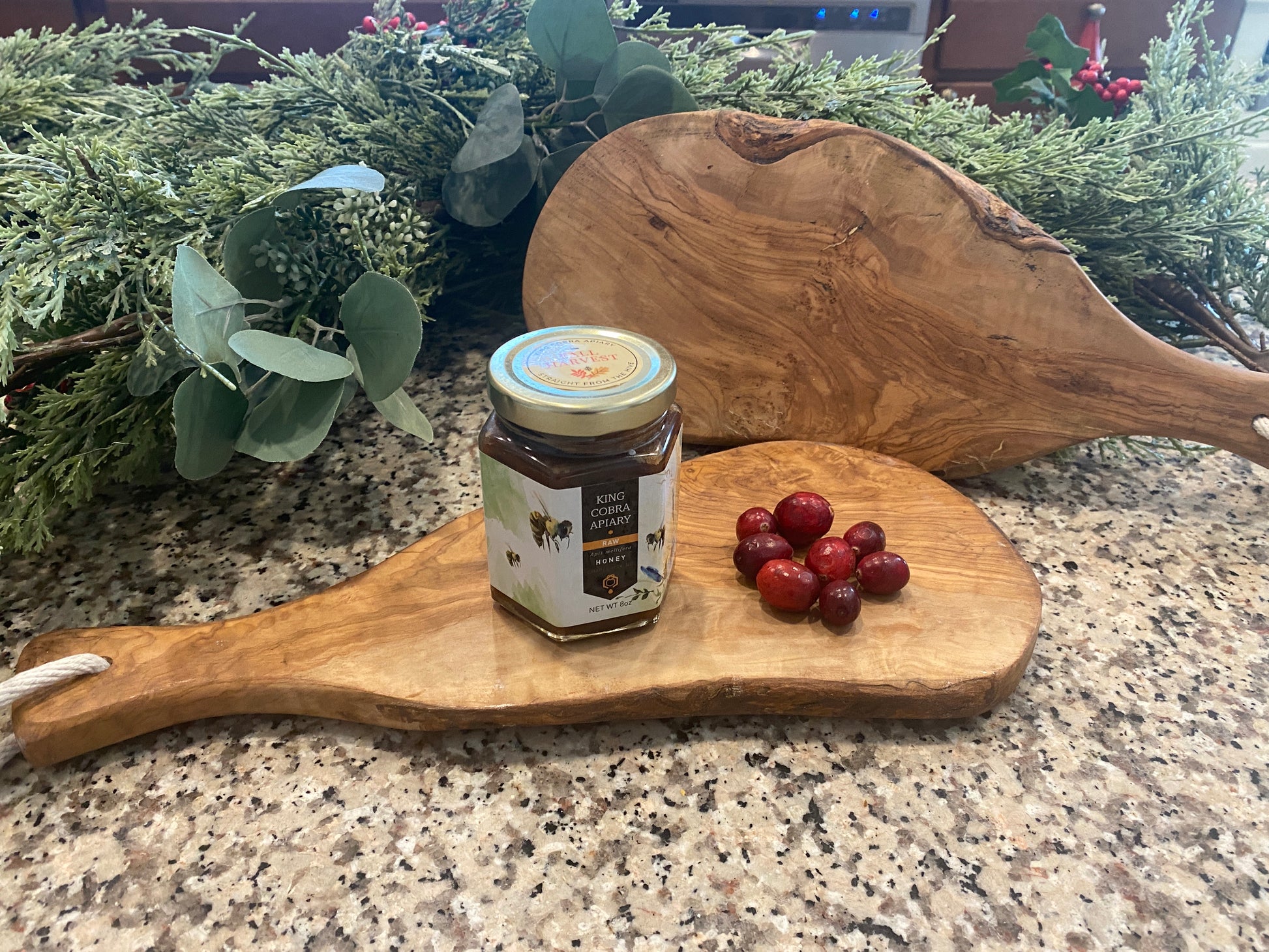 Two live edge wooden cutting boards with organic, irregular shapes are displayed on a granite countertop. One board lies flat holding a jar of honey and a small cluster of red grapes, while the other leans upright behind it. A greenery garland with eucalyptus and berries sits in the background, emphasizing the boards’ natural wood grain and rustic, handcrafted look.