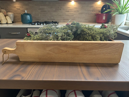 A long rectangular wooden cutting board sits on a darker wood countertop, styled on a kitchen island. Behind it, a festive greenery garland with red berries adds a seasonal touch with a modern stone kitchen backsplash and counter. The board’s natural wood grain and elongated shape make it ideal for serving charcuterie or entertaining displays.