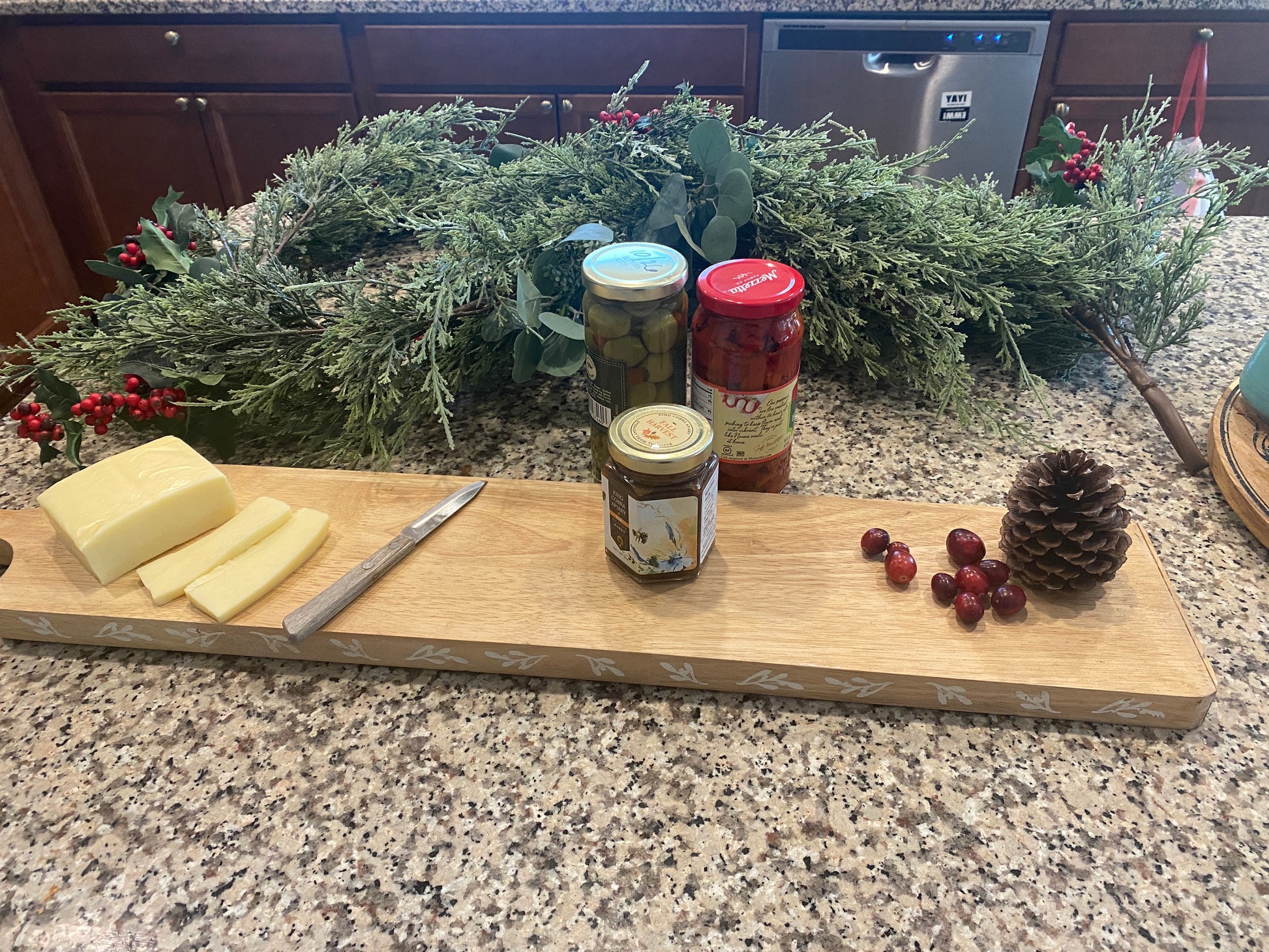 A long rectangular wooden cutting board sits on a granite countertop, styled with sliced cheese, a knife, jars of olives and preserves, and fresh cranberries. Behind it, a festive greenery garland with red berries adds a seasonal touch. The board’s natural wood grain and elongated shape make it ideal for serving charcuterie or entertaining displays.