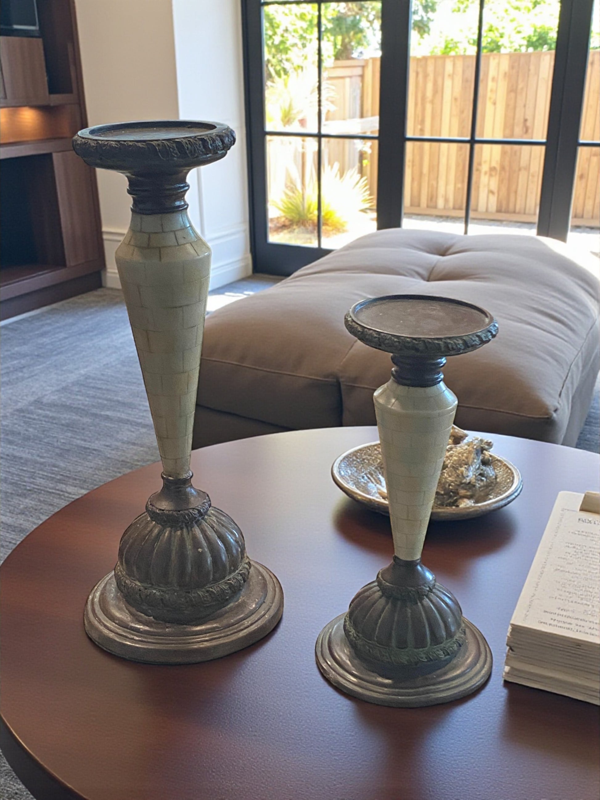 Two decorative candle holders on a table with a couch and window in the background.  Two marble and metal candle holders with a tapered design and aged patina sit on a darker wood  tabletop in a living room. The bases feature dark metal detailing, while the stems showcase light marble accents. Sunlight from nearby windows highlights their texture, with ottoman and fireplace in the background adding a natural touch, creating a warm, elegant, and timeless setting.
