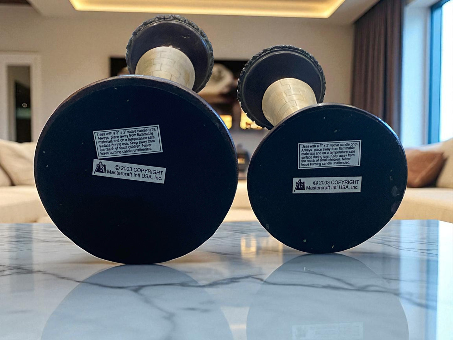 The bottom of two marble and metal candle holders, showing the makers information.  A tapered design and aged patina sit on a stone tabletop. The bases feature dark metal detailing, while the stems showcase light marble accents. Sunlight from nearby windows highlights their texture, with a modern living room background adding a natural touch, creating a warm, elegant, and timeless setting.