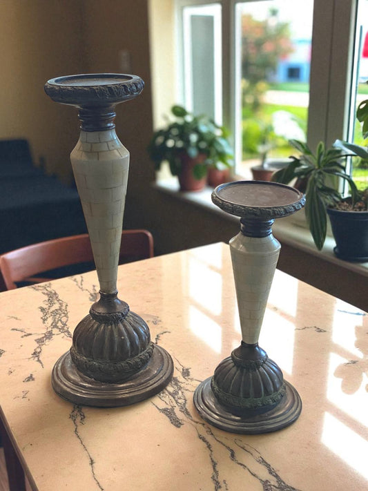Two marble and metal candle holders with a tapered design and aged patina sit on a stone tabletop. The bases feature dark metal detailing, while the stems showcase light marble accents. Sunlight from nearby windows highlights their texture, with plants in the background adding a natural touch, creating a warm, elegant, and timeless setting.