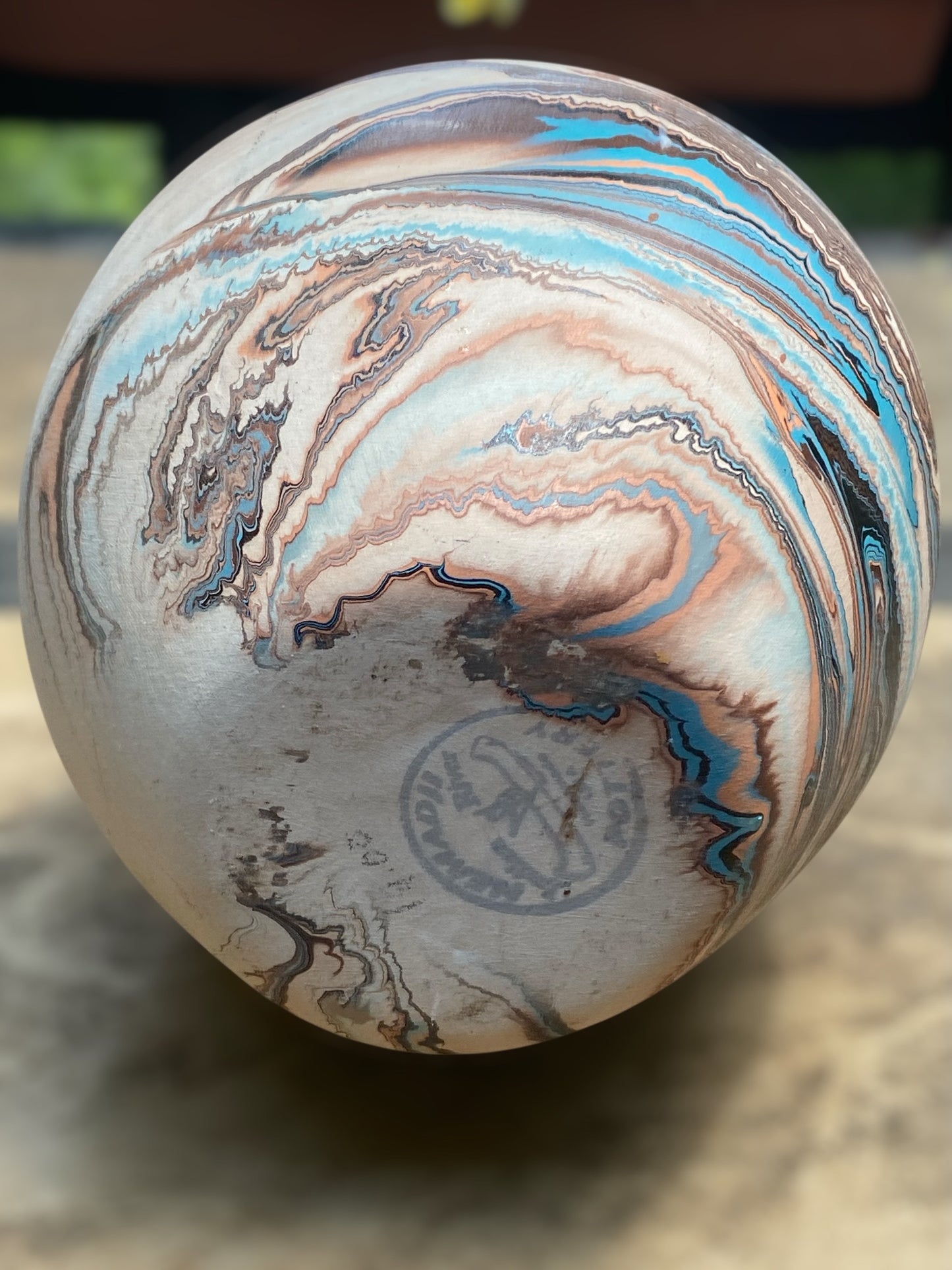 Close up of the bottom makers mark on a small Southwest-style ceramic vase with a rounded shape and narrow opening sits on a light stone surface. The vase features marbled layers of cream, rust, and subtle blue tones resembling desert landscapes. In the softly blurred background, green succulents in white pots sit near a window, creating a calm, natural setting that highlights the vase’s earthy, handcrafted appearance. Great for bohemian, earthy, or rustic decor.  