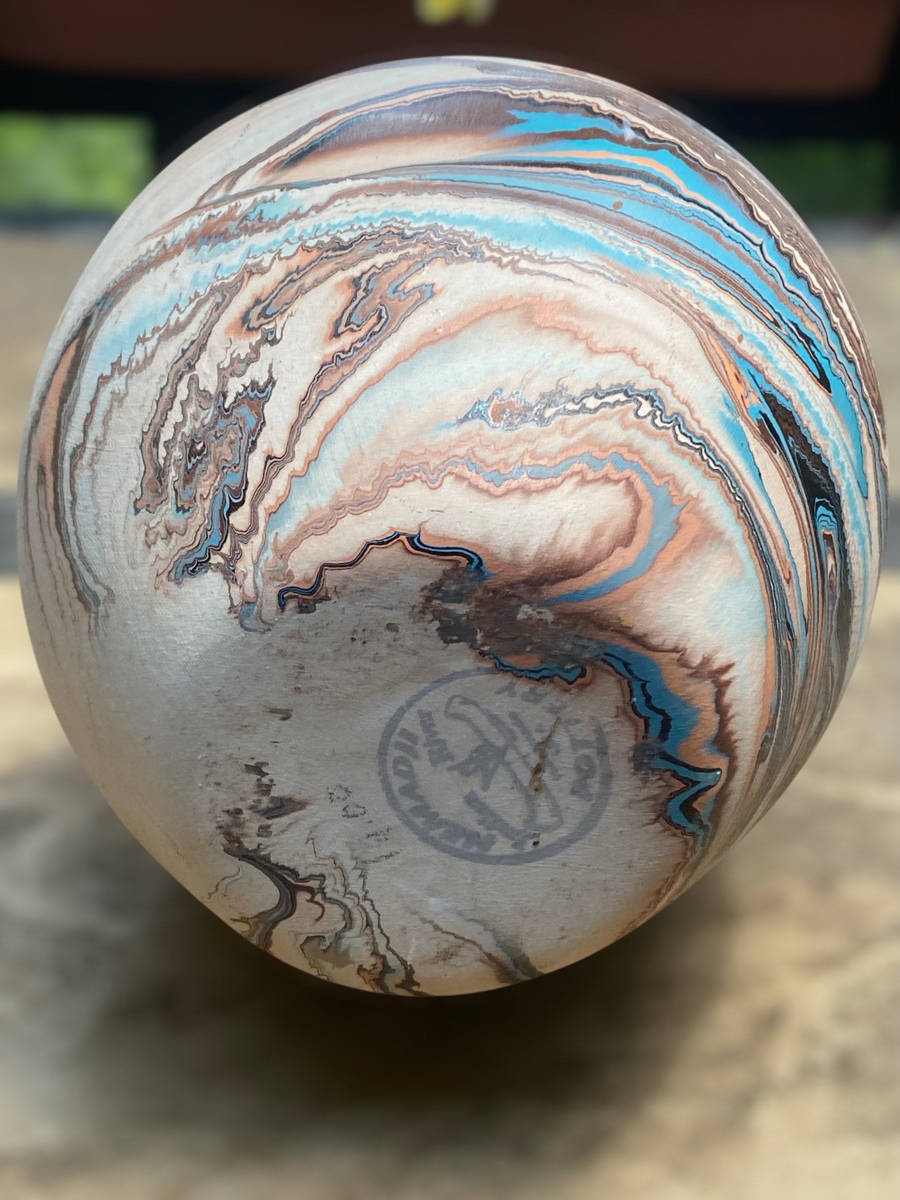 Close up of the bottom makers mark on a small Southwest-style ceramic vase with a rounded shape and narrow opening sits on a light stone surface. The vase features marbled layers of cream, rust, and subtle blue tones resembling desert landscapes. In the softly blurred background, green succulents in white pots sit near a window, creating a calm, natural setting that highlights the vase’s earthy, handcrafted appearance. Great for bohemian, earthy, or rustic decor.  