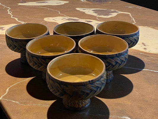 A set of six ceramic bowls with a textured, woven-style exterior and warm glazed interior are arranged on a brown marble tabletop. Each bowl sits on a small pedestal base, adding height and visual interest. The earthy tones and handcrafted look create a cozy, vintage-inspired feel, suitable for serving food or use as decorative home accents.  Hawaiian decor.    