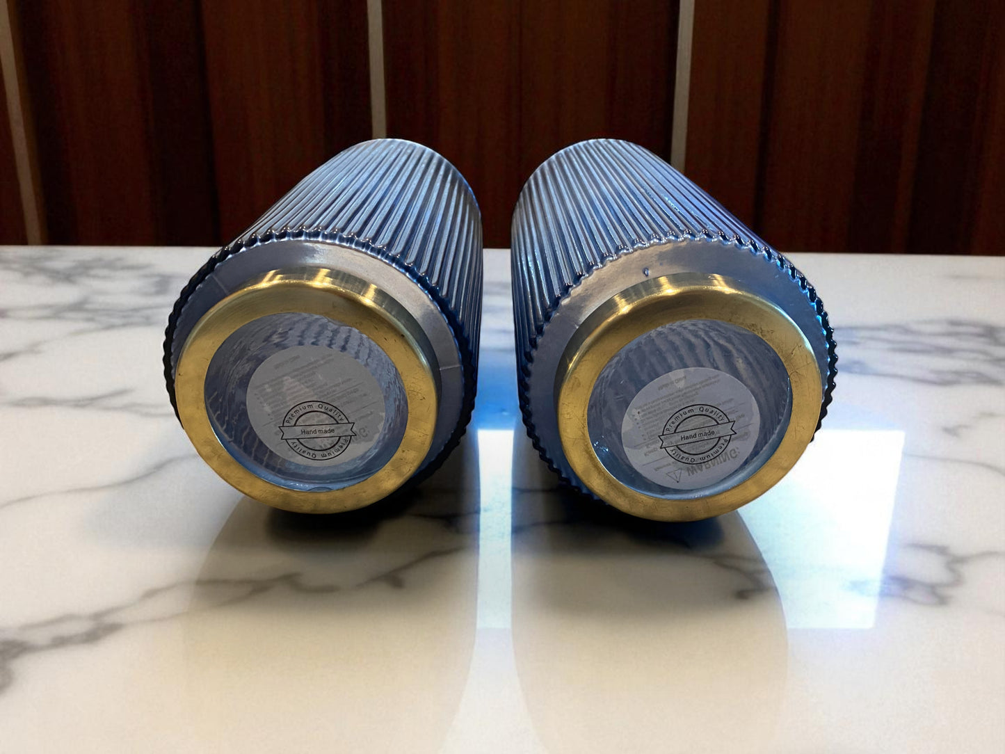 Two blue and gold cylindrical objects on a marble surface with a wooden background.  Two matching blue fluted glass vases , laying on their sides to see the bottom showing the makers mark, with a ribbed texture sit on a marble kitchen island. The deep blue color and vertical lines create a sleek, elegant look. Wooden cabinets are visible in the background, adding warmth and context to the contemporary setting.