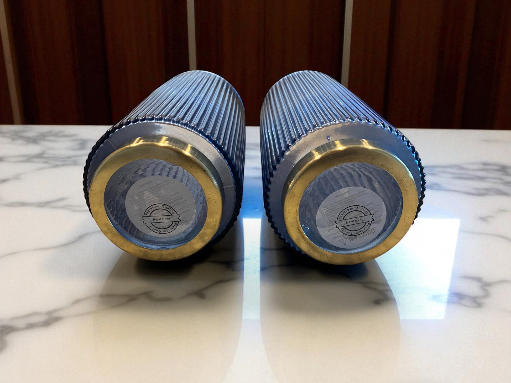 Two blue and gold cylindrical objects on a marble surface with a wooden background.  Two matching blue fluted glass vases , laying on their sides to see the bottom showing the makers mark, with a ribbed texture sit on a marble kitchen island. The deep blue color and vertical lines create a sleek, elegant look. Wooden cabinets are visible in the background, adding warmth and context to the contemporary setting.