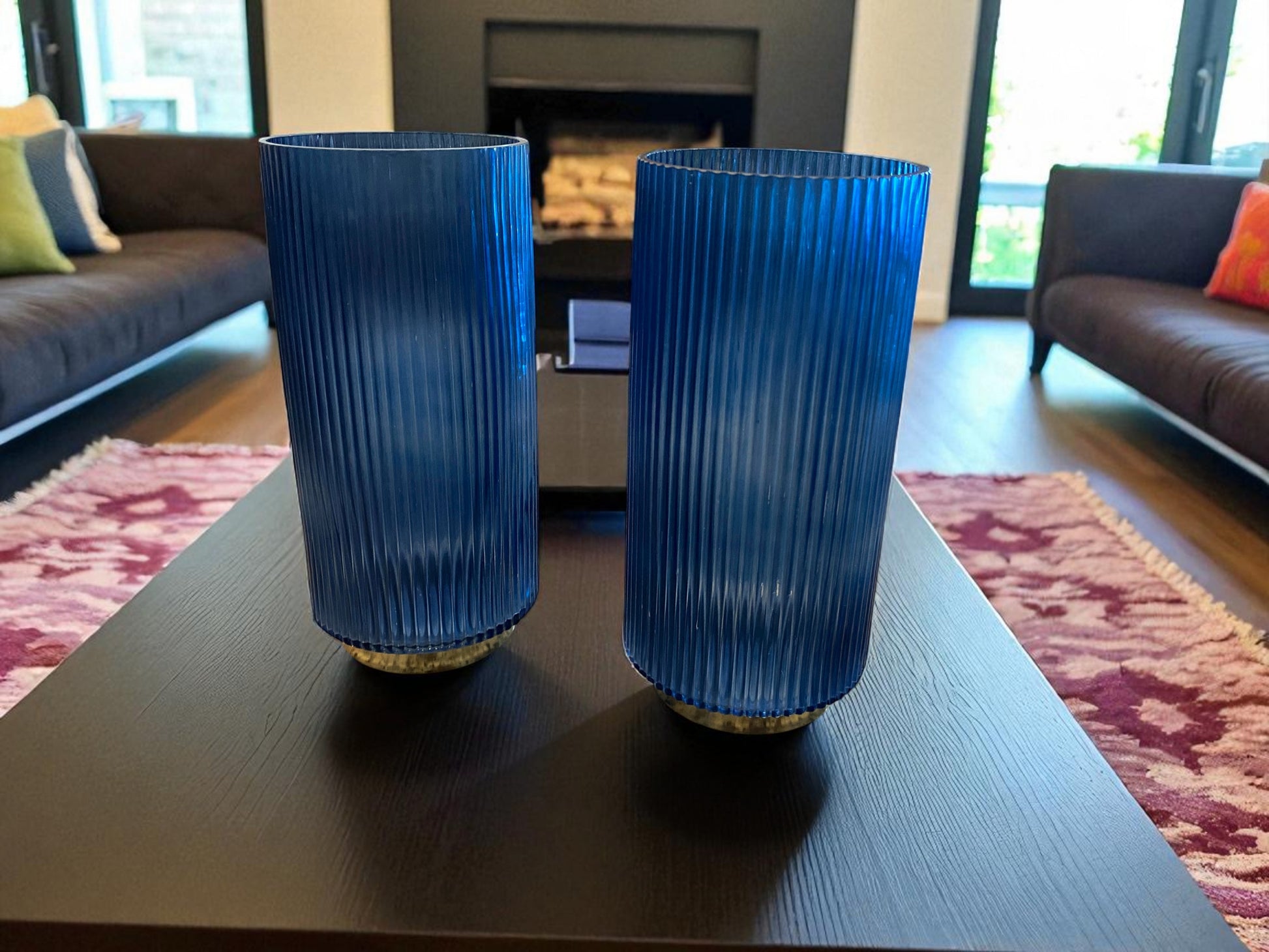 Two matching blue fluted glass vases with a ribbed texture sit on a dark coffee table in a modern living room. The deep blue color and vertical lines create a sleek, elegant look. Sofas, a fireplace, and a patterned rug are visible in the background, adding warmth and context to the contemporary setting.
