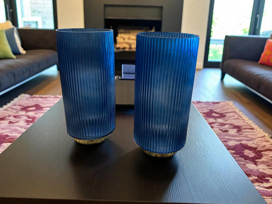Two matching blue fluted glass vases with a ribbed texture sit on a dark coffee table in a modern living room. The deep blue color and vertical lines create a sleek, elegant look. Sofas, a fireplace, and a patterned rug are visible in the background, adding warmth and context to the contemporary setting.