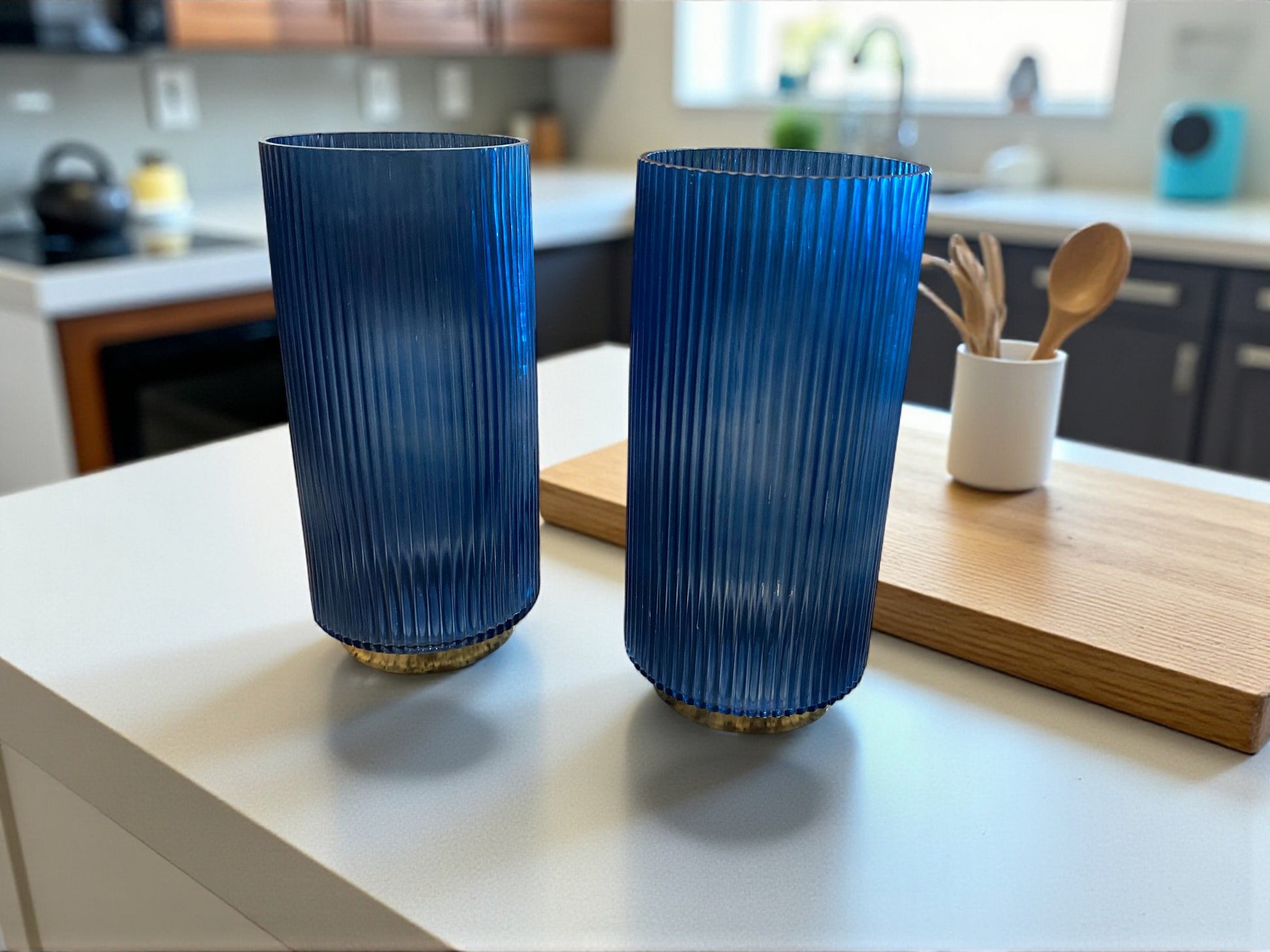 Two matching blue fluted glass vases,  with a ribbed texture sit on a white modern kitchen island. The deep blue color and vertical lines create a sleek, elegant look. Colorful kitchen with window, cutting board, and utensil holder are visible in the background, adding warmth and context to the contemporary setting.
