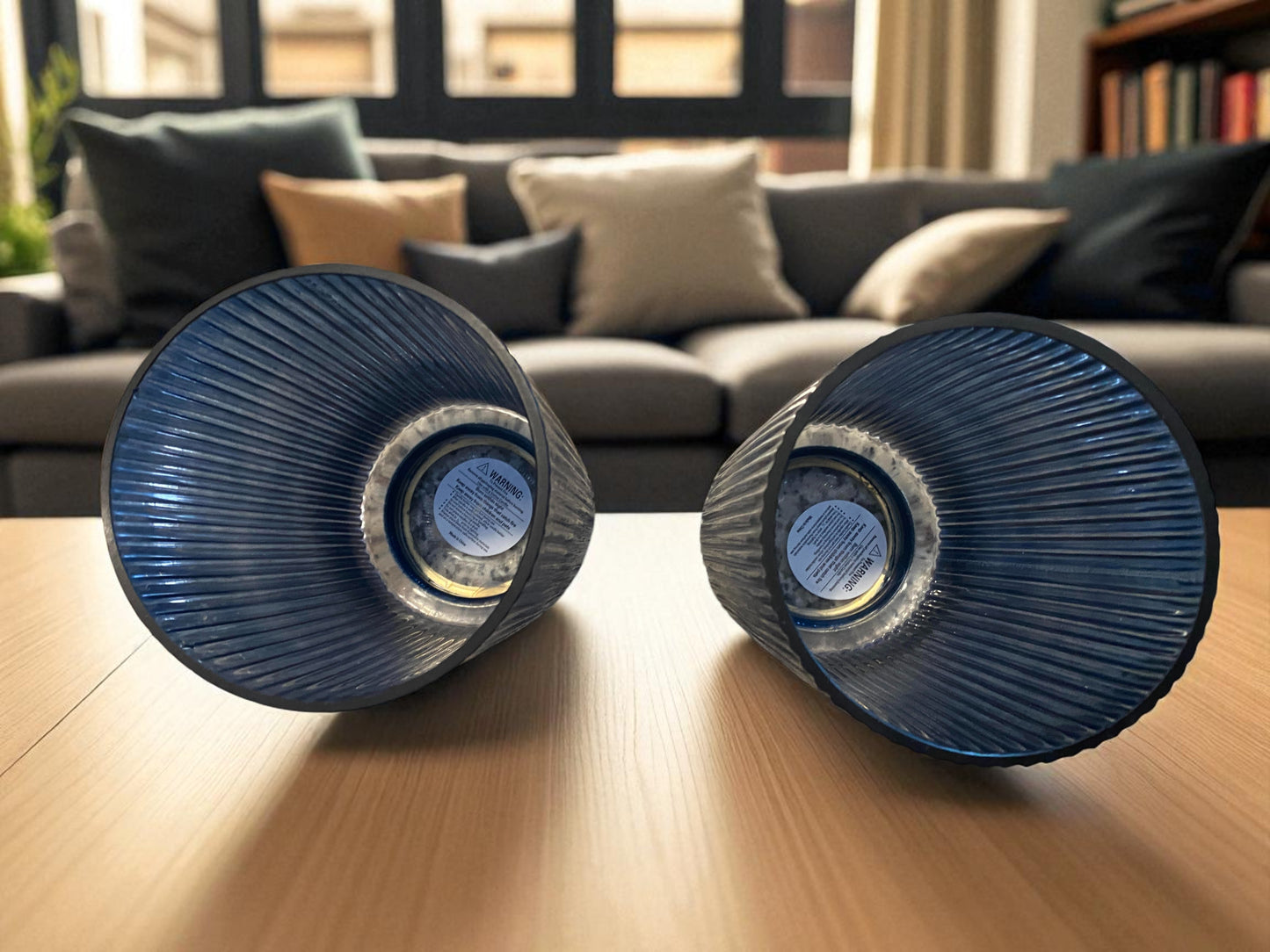 Two matching blue fluted glass vases , laying on their sides, with a ribbed texture sit on a coffee table in a modern living room. The deep blue color and vertical lines create a sleek, elegant look. Sofas, with pillows, a widow, and bookcase are visible in the background, adding warmth and context to the contemporary setting.