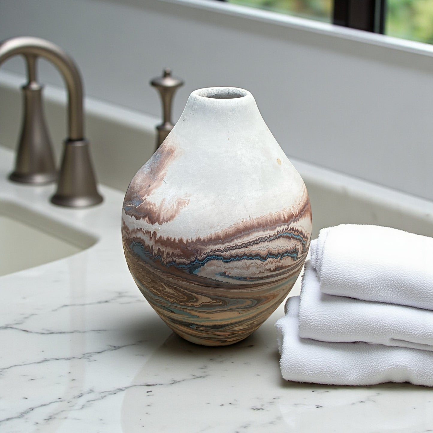A small Southwest-style ceramic vase with a rounded shape and narrow opening sits on a light stone surface. The vase features marbled layers of cream, rust, and subtle blue tones resembling desert landscapes. In the softly blurred background, modern bathroom sink near a window and white towels in the foreground, creating a calm, natural setting that highlights the vase’s earthy, handcrafted appearance. Great for bohemian, earthy, or rustic decor.  
