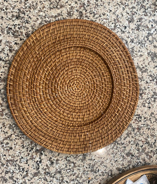 A round wicker charger plate with a tightly woven rattan design is displayed on a speckled stone countertop. The charger features a warm natural brown tone and a circular pattern that radiates from the center.  Great for bohemian, rustic, or bringing warmth to a modern dining table.  