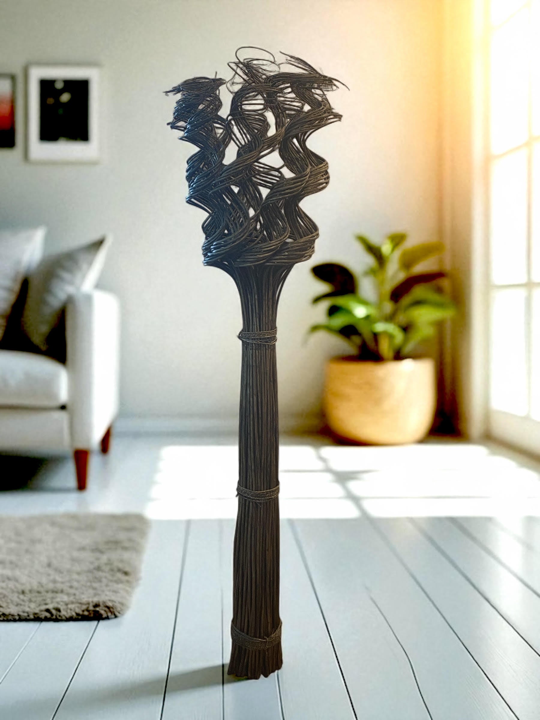 A tall floor-standing wicker sculpture made of bundled natural fibers stands in a warm, modern living room. The base is tightly bound, extending upward into an intricate, twisted woven design that creates an abstract, sculptural form. It is placed near a large window with natural light, a modern couch and large planter in the background, highlighting its organic texture and vertical presence.