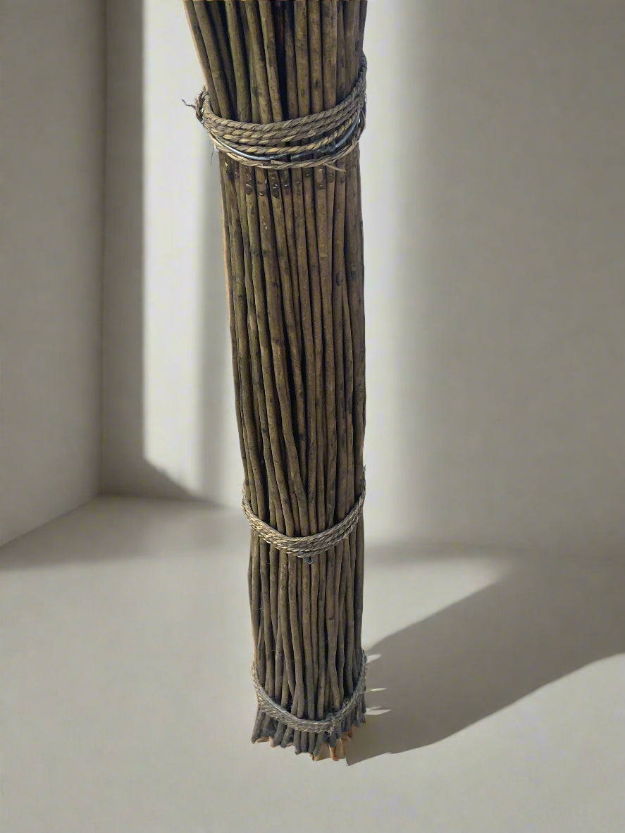 A close up of the base of a tall floor-standing wicker sculpture made of bundled natural fibers stands in a minimalist room. The base is tightly bound, extending upward into an intricate, twisted woven design that creates an abstract, sculptural form. It is placed near a large window with natural light, highlighting its organic texture and vertical presence.