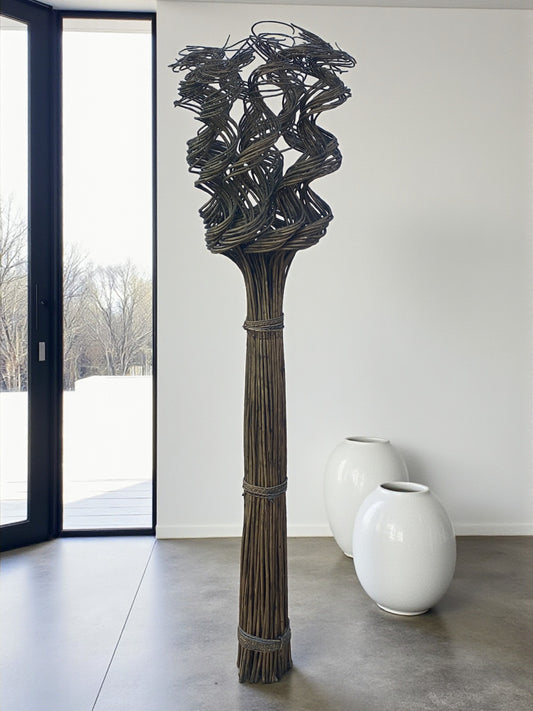 A tall floor-standing wicker sculpture made of bundled natural fibers stands in a minimalist room. The base is tightly bound, extending upward into an intricate, twisted woven design that creates an abstract, sculptural form. It is placed near a large window with natural light, with two smooth white ceramic floor vases in the background, highlighting its organic texture and vertical presence.
