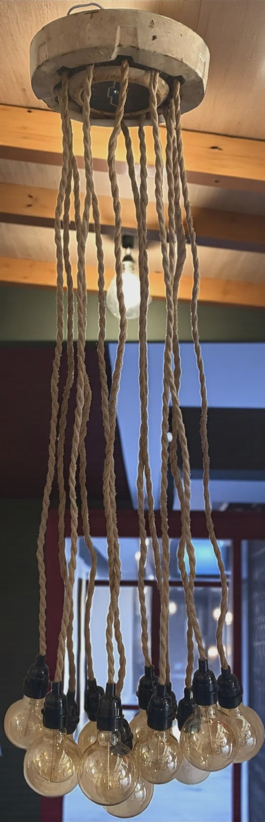 A unique, vintage-inspired chandelier made from a Chevy hubcap as ceiling cover and is mounted to a wood ceiling, with multiple twisted rope electrical cords hanging down. Each cord holds an exposed Edison bulb with a warm amber glow. The fixture has an industrial, rustic look and is shown in a modern interior space, highlighting its unique design and vintage automotive influence.