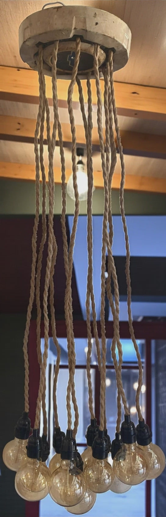 A unique, vintage-inspired chandelier made from a Chevy hubcap as ceiling cover and is mounted to a wood ceiling, with multiple twisted rope electrical cords hanging down. Each cord holds an exposed Edison bulb with a warm amber glow. The fixture has an industrial, rustic look and is shown in a modern interior space, highlighting its unique design and vintage automotive influence.