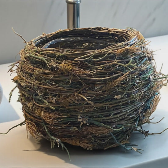 A bird’s nest-style planter made of intertwined mixture of artificial twigs and natural fibers sits on a bathroom countertop, with a towel and glass container next to it. The organic, textured design features earthy brown and green tones, adding a rustic, nature-inspired touch. The soft background of a white/gray marbled modern bathroom creates a warm, balanced contrast between natural materials and clean, contemporary surfaces.  Great for any room of a modern home - kitchen, bathroom, office, den, or foyer